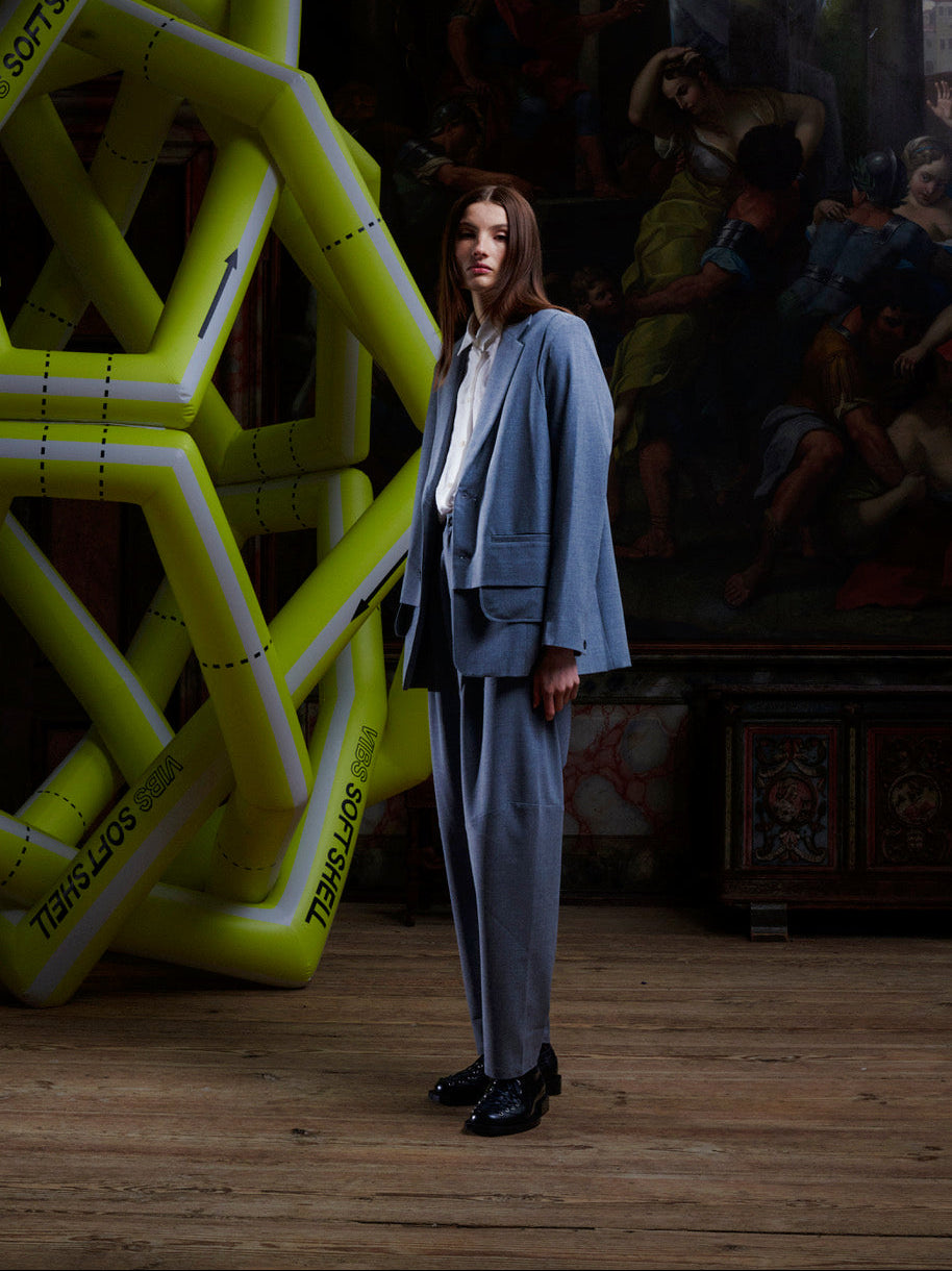 Full-body shot of model in grey Henrik Vibskov pants paired with matching blazer, standing in artistic indoor environment with geometric installation