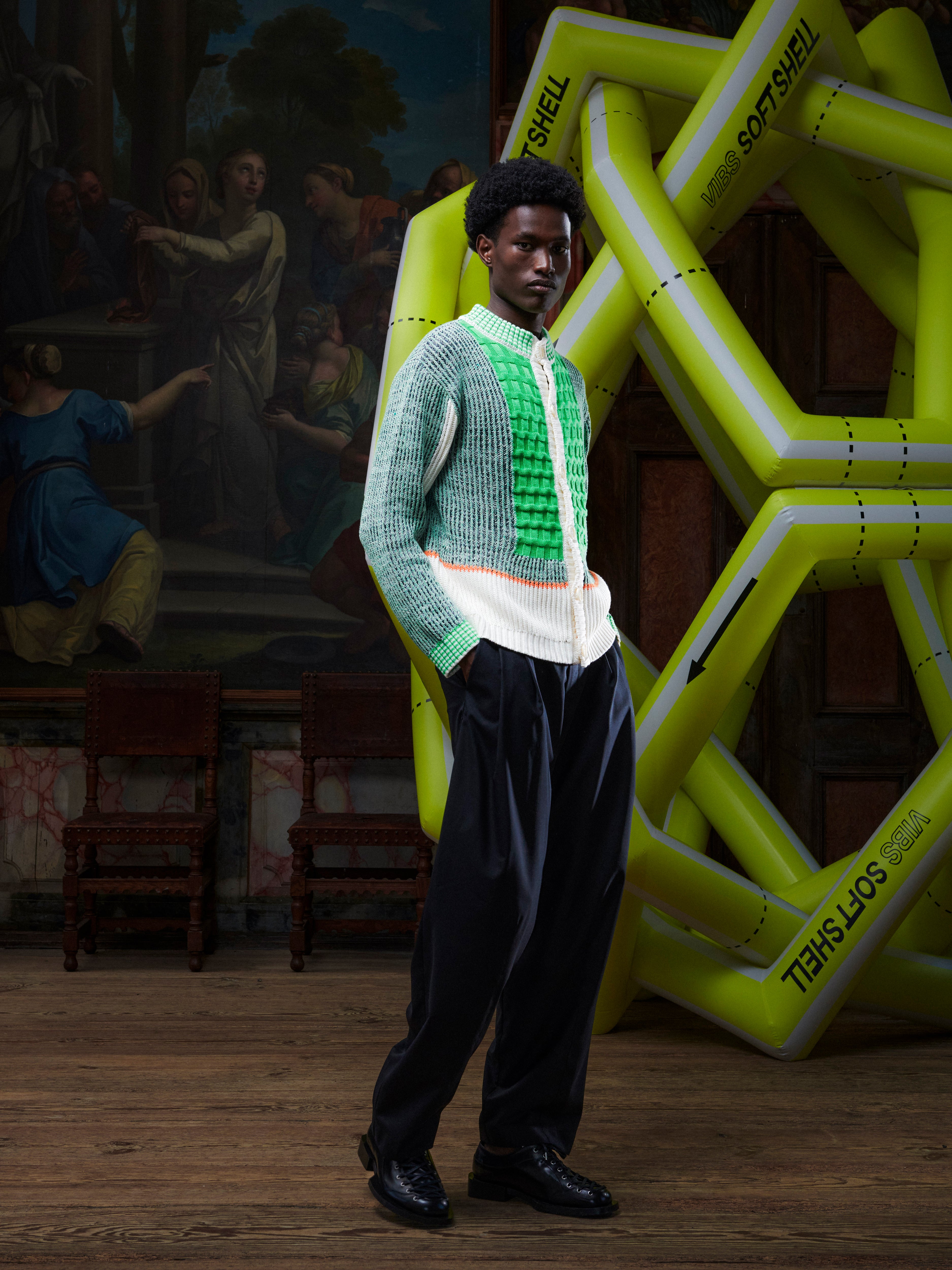 Side view of the male model wearing the Abundance Cardigan from Henrik Vibskov Spring/Summer 2026 Studio Collection, showing the fit of the long sleeves and the green, grey, and navy block pattern with beige detailing.