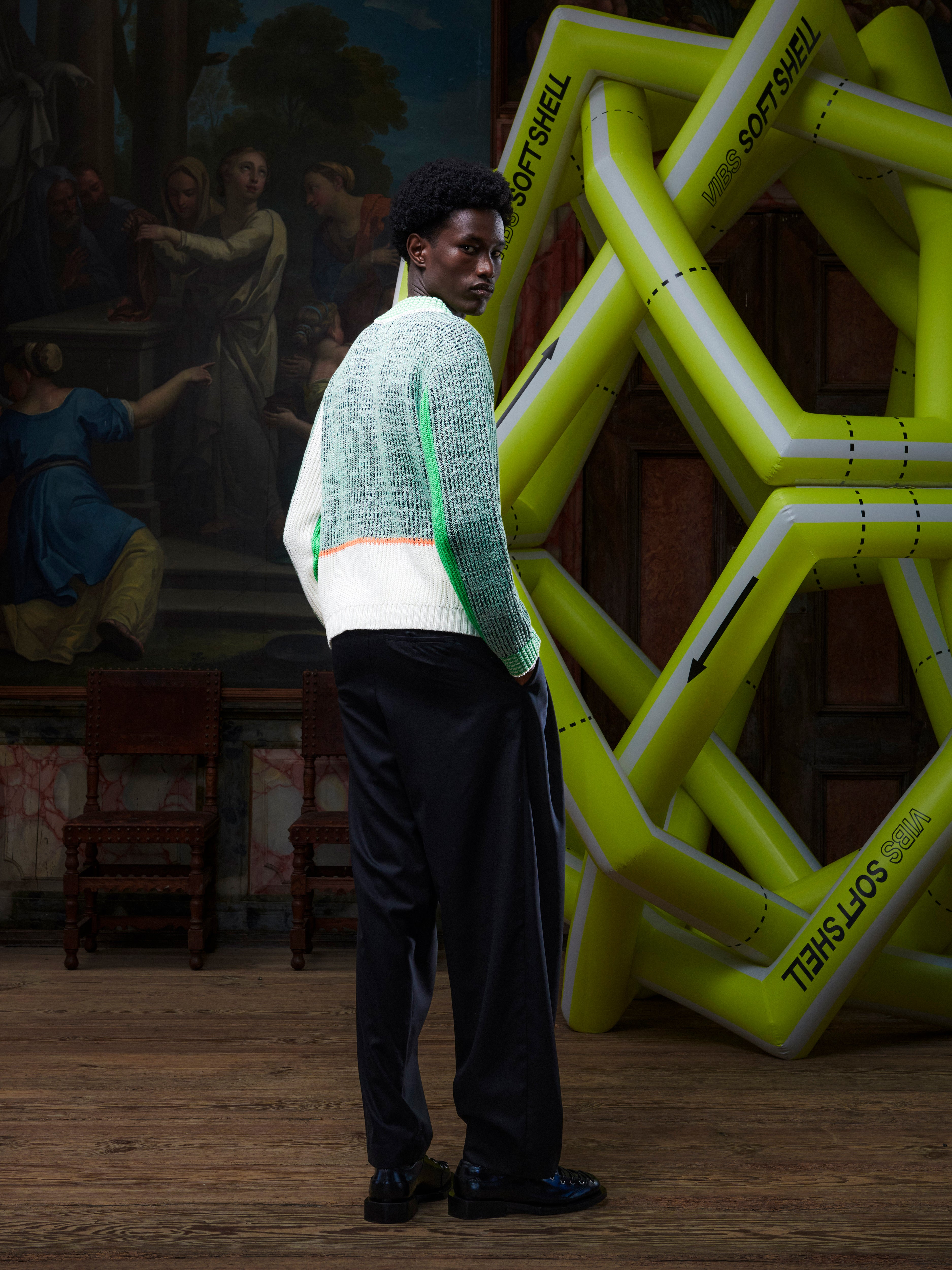 Back view of the male model wearing the Abundance Cardigan from Henrik Vibskov Spring/Summer 2026 Studio Collection, highlighting the green, grey, and navy block-color design and straight hem.