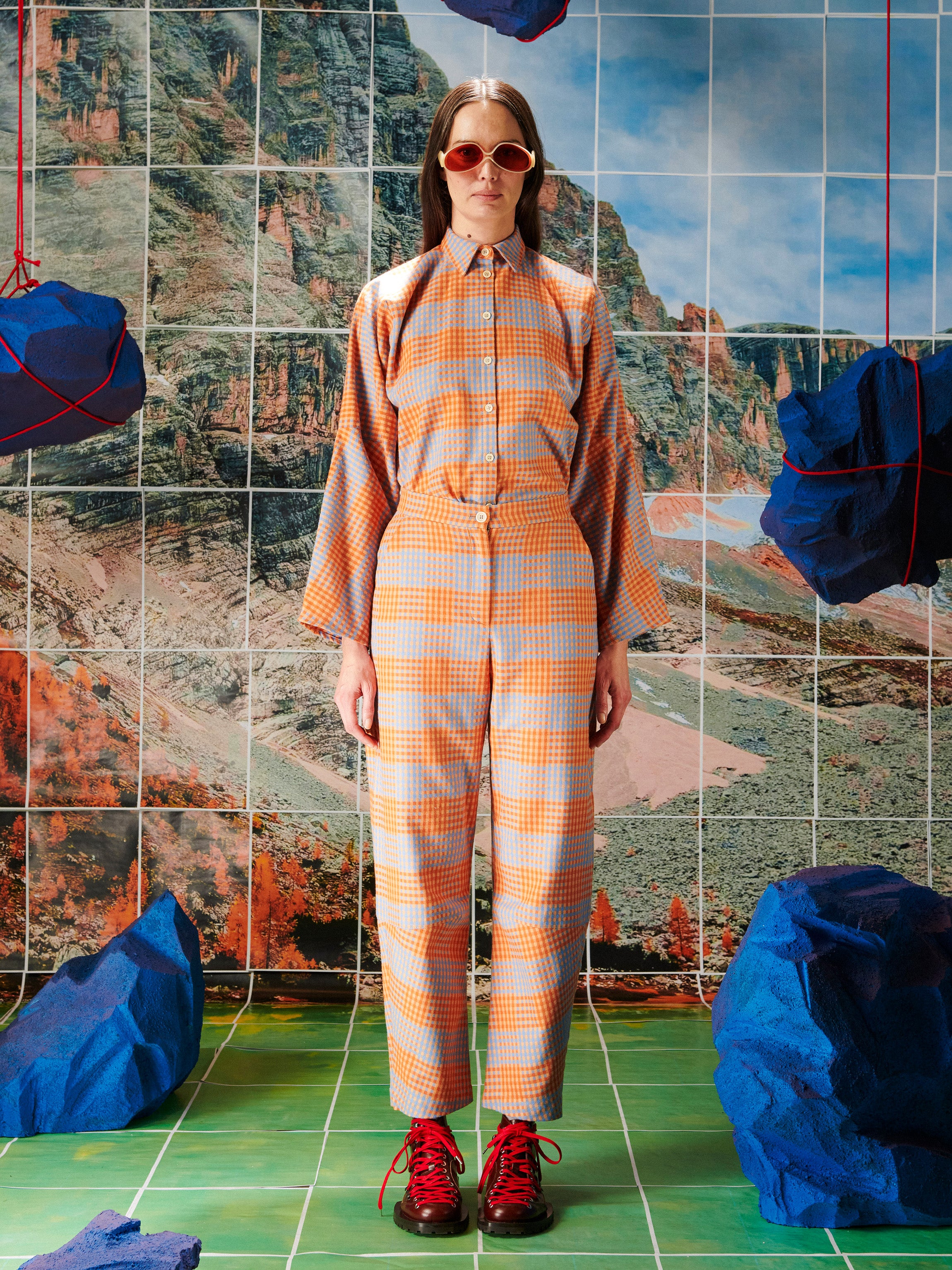 Woman in a plaid outfit standing in front of a tiled wall with mountain and blue rock designs.