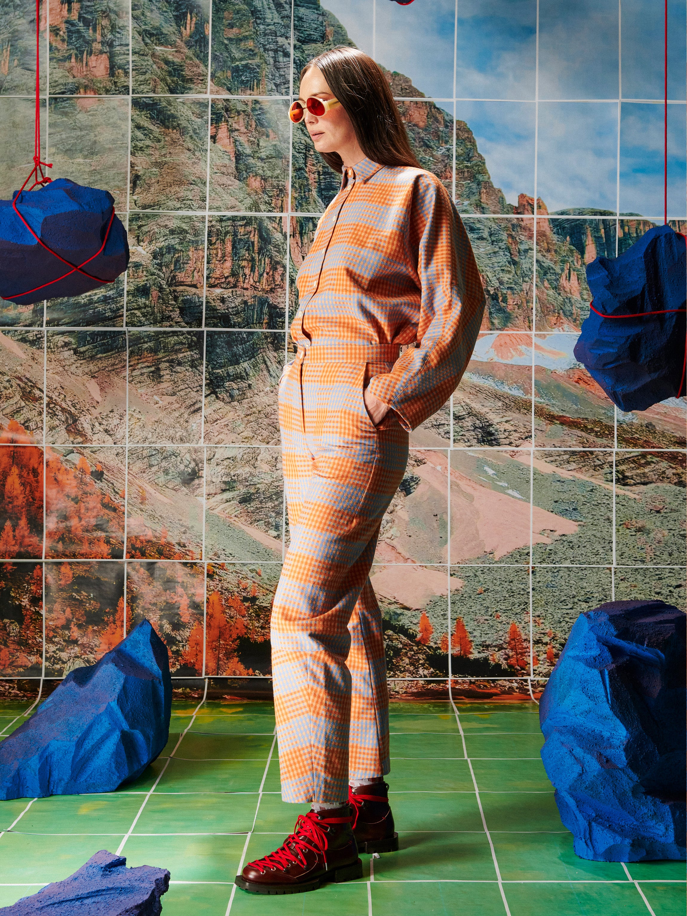 Person wearing an orange and gray striped outfit standing in front of a tiled wall with a mountainous landscape.