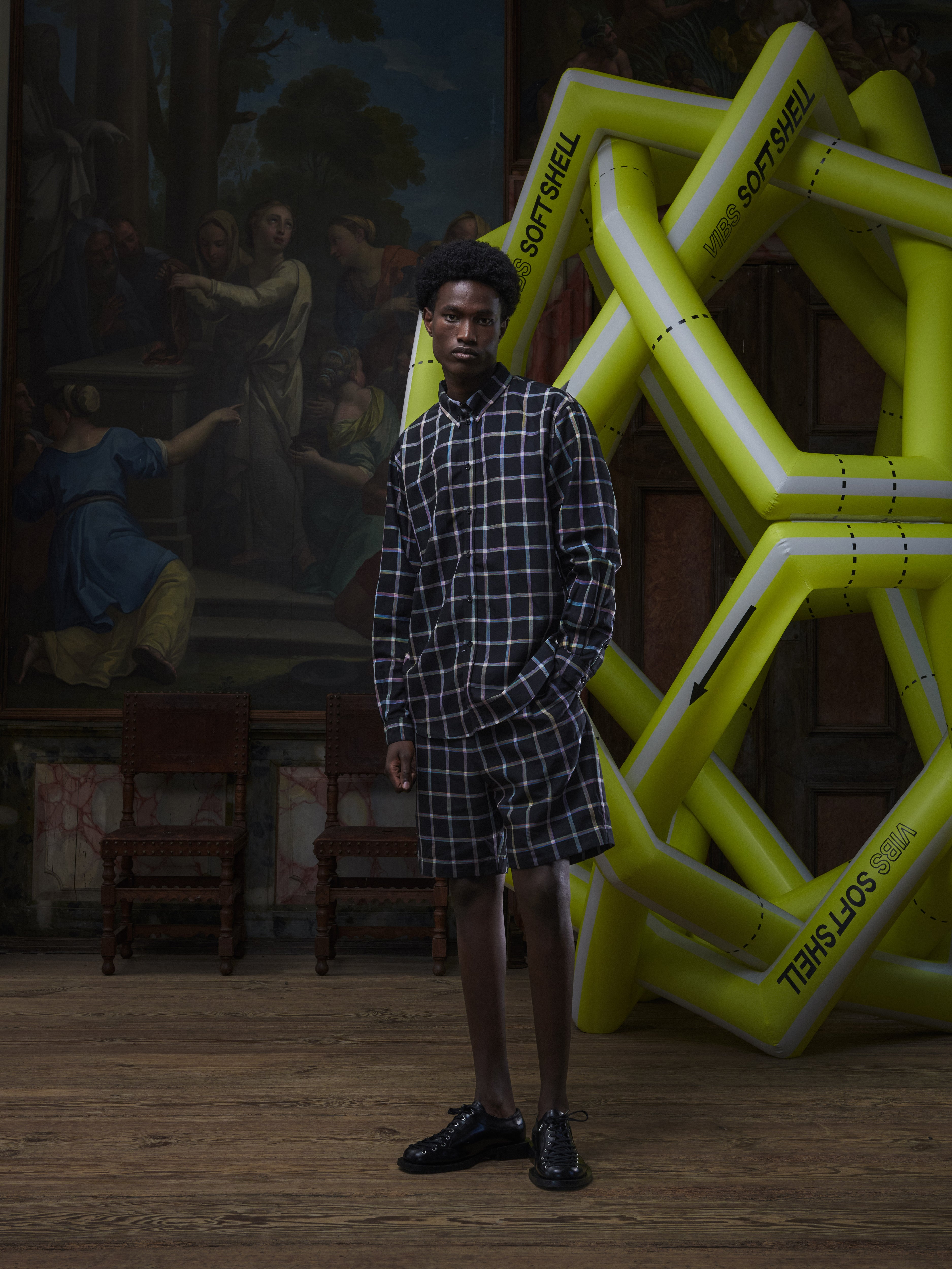 Front view of the male model wearing the Block Shirt in Black Checks from Henrik Vibskov Spring/Summer 2026 Studio Collection, showing the colourful check pattern, long sleeves, and front button closure.