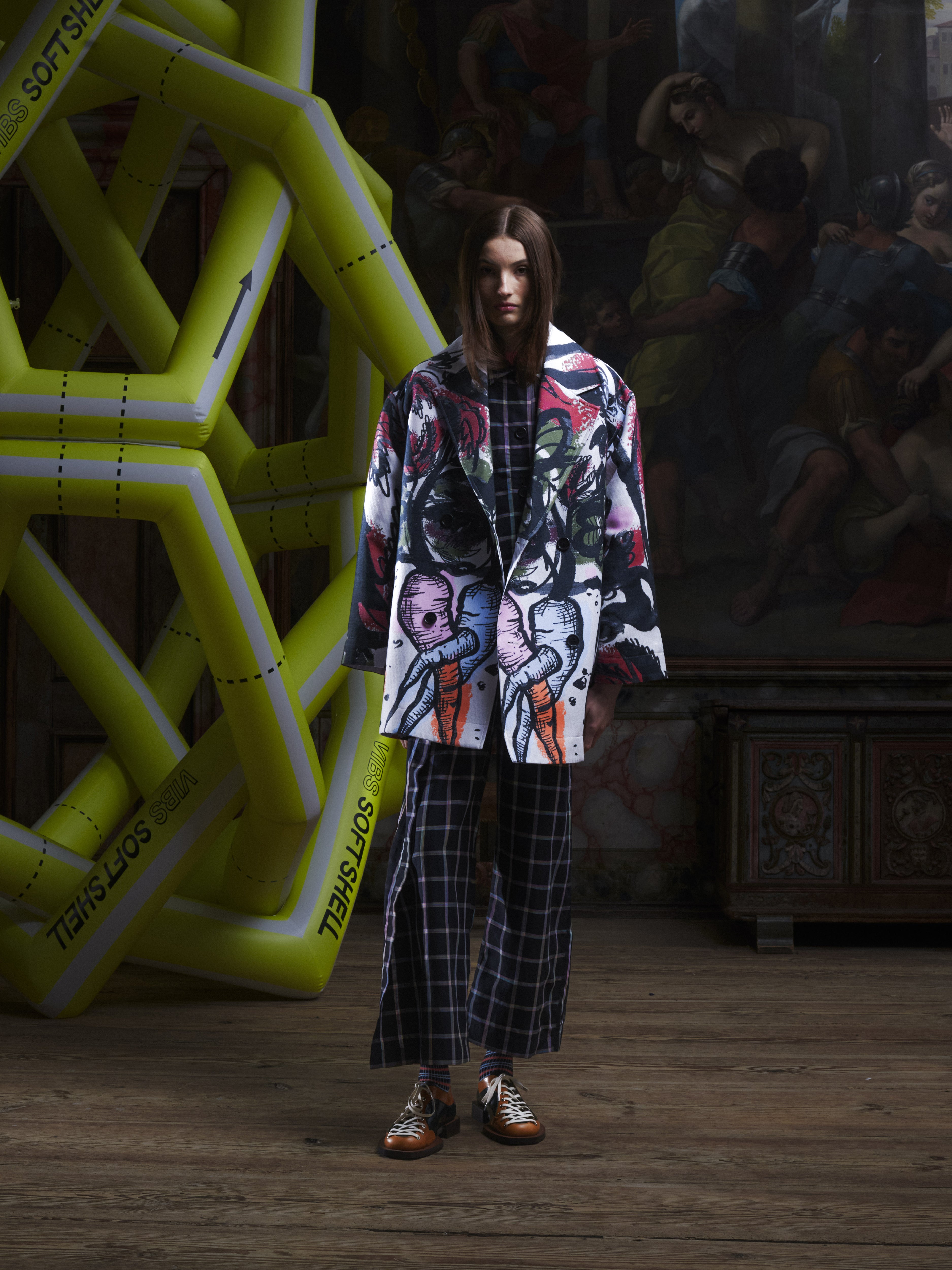 Front view of the female model wearing the Care Coat in Carrots from Henrik Vibskov Spring/Summer 2026 Studio Collection, showing the multicoloured carrot print, double-breasted closure, long sleeves, and pockets.