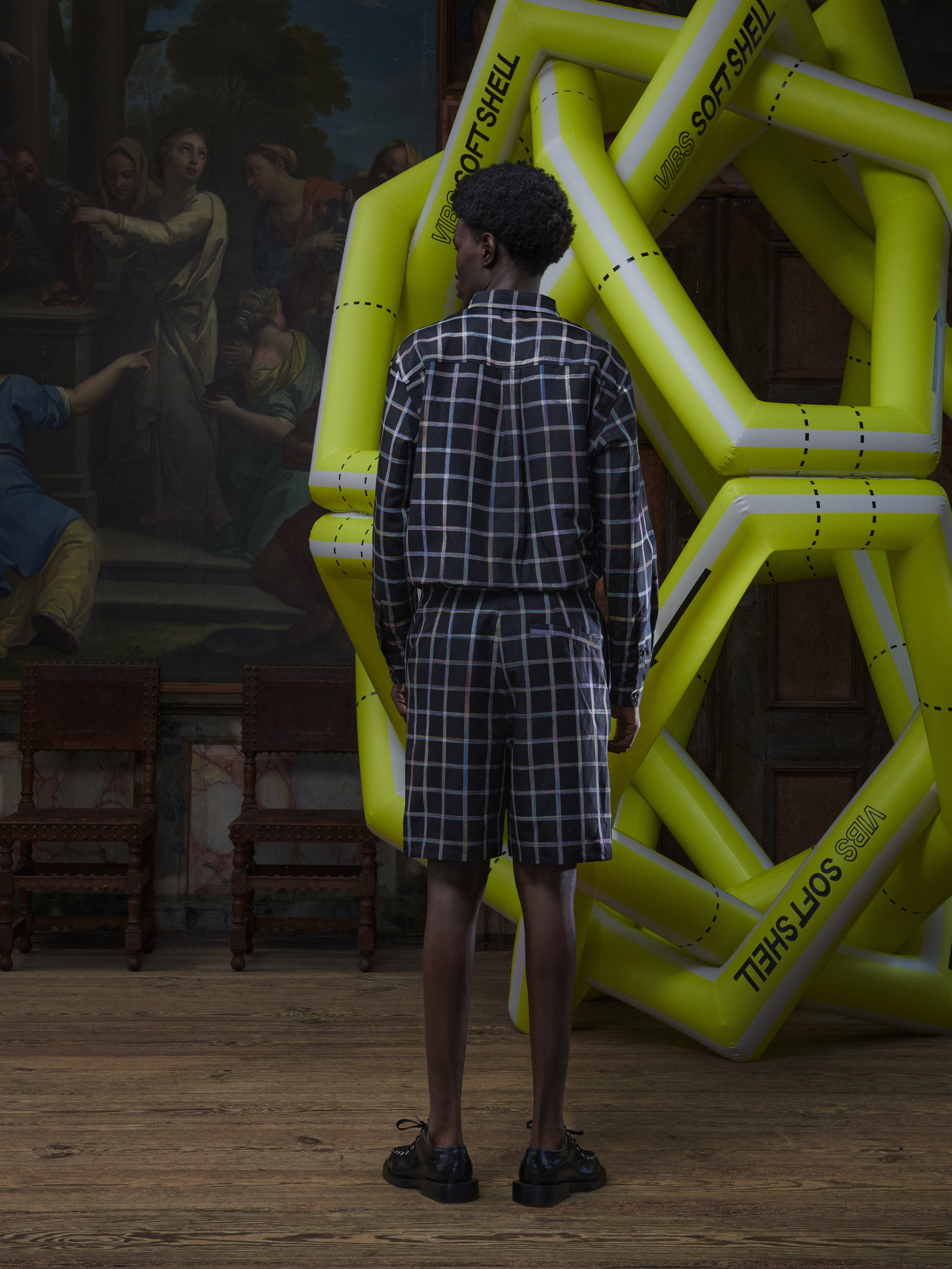 Back full-body view of a male model wearing the Claus Shorts in Black Checks from Henrik Vibskov Spring/Summer 2026 Studio Collection, showing the back silhouette, belt loops, and straight leg opening.
