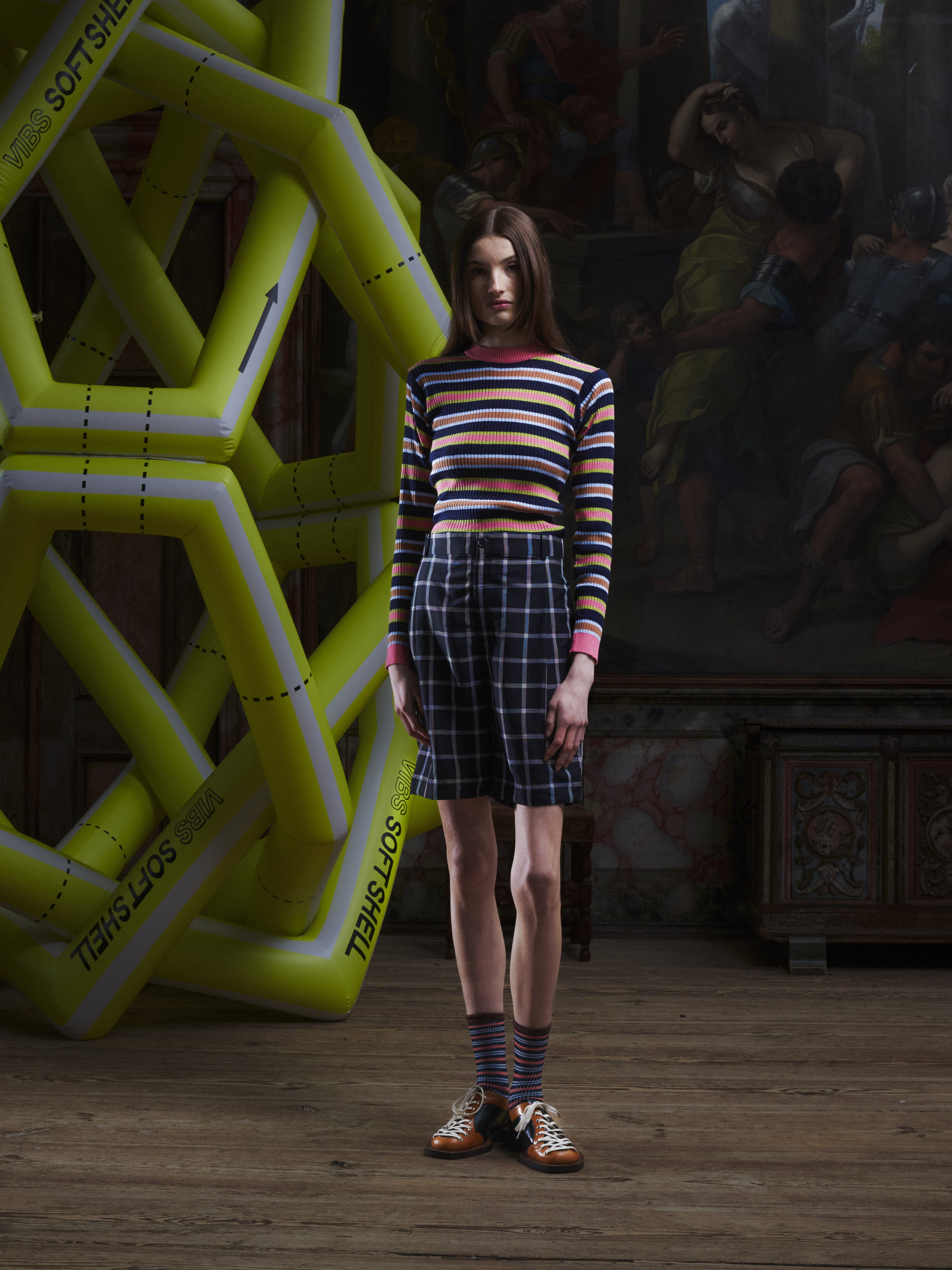 Front view of a female model wearing the Claus Shorts in Black Checks from Henrik Vibskov Spring/Summer 2026 Studio Collection, showing the multicoloured check pattern, front closure, and unisex fit.