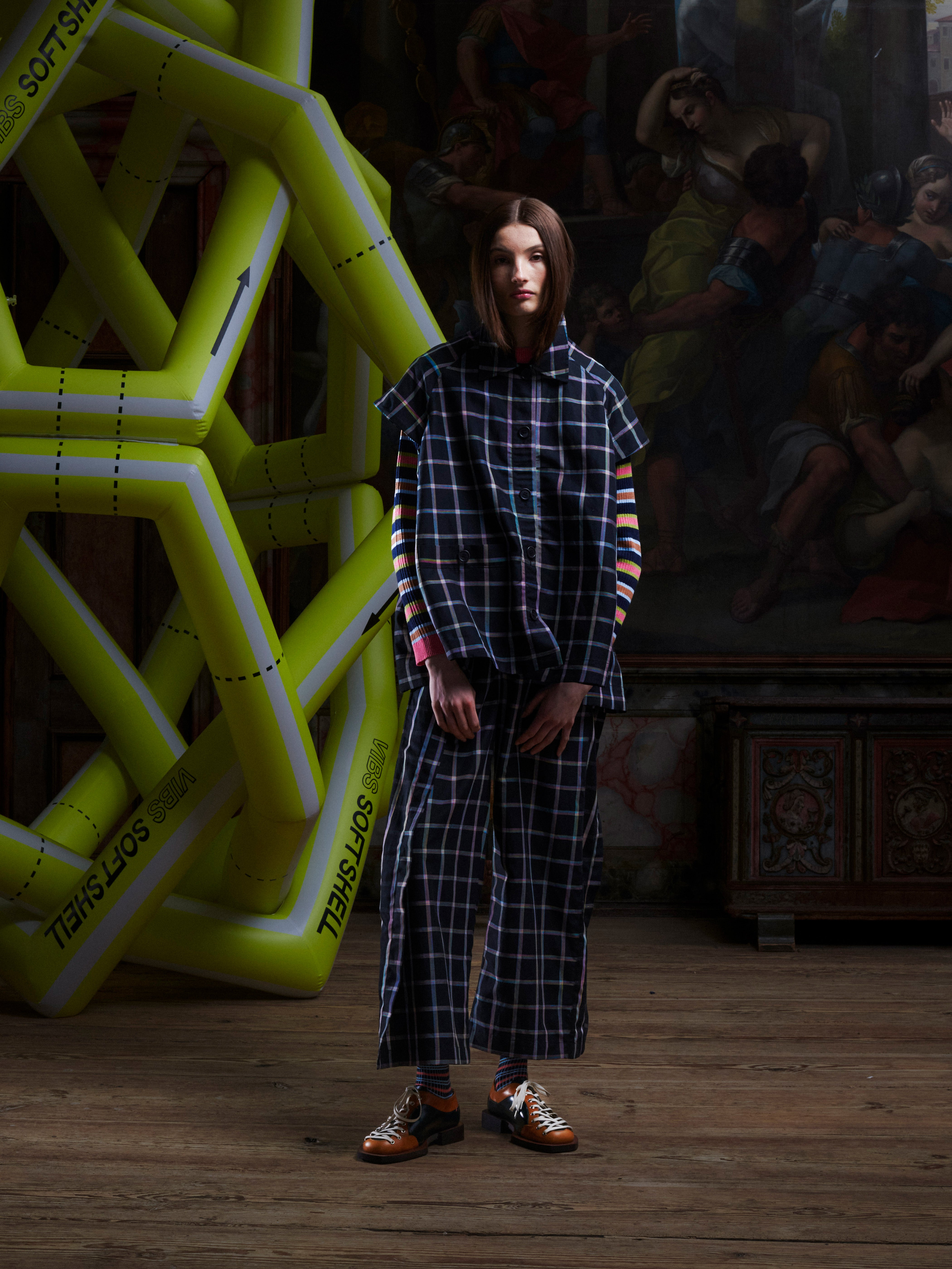 Front view of a female model wearing the Cover Coat in Black Checks from Henrik Vibskov Spring/Summer 2026 Studio Collection with the sleeves detached, showing the sleeveless configuration, collar, front button closure, and check-patterned fabric.