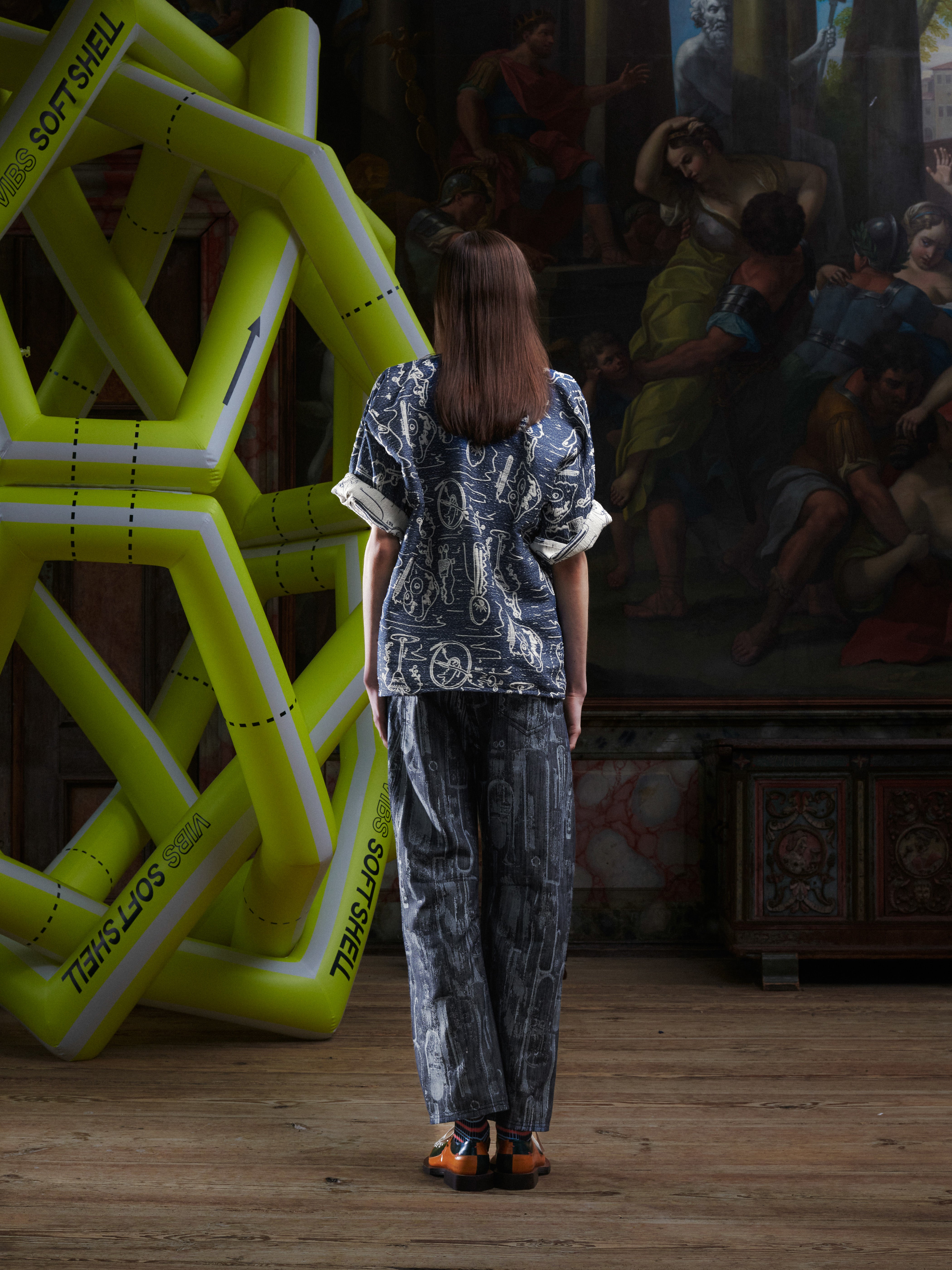 Back view of the female model wearing the grey-blue Dart Polo Shirt, showing the full doodle print and relaxed oversized fit. Model is 185cm and wears size L.