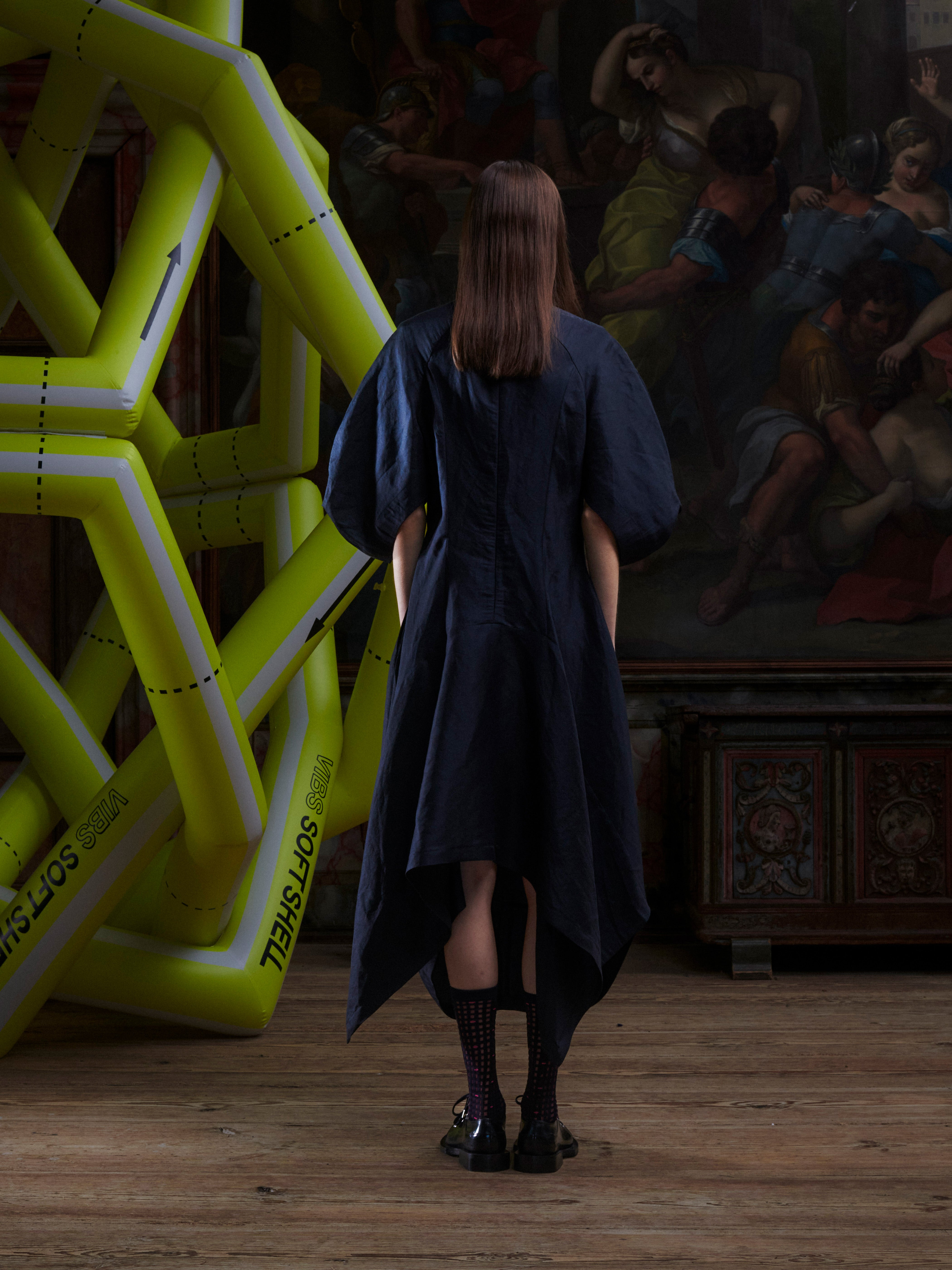 Back view of model wearing the Double Bass Dress in Black, showing the clean black design, hourglass structure, and asymmetric low–high hem.