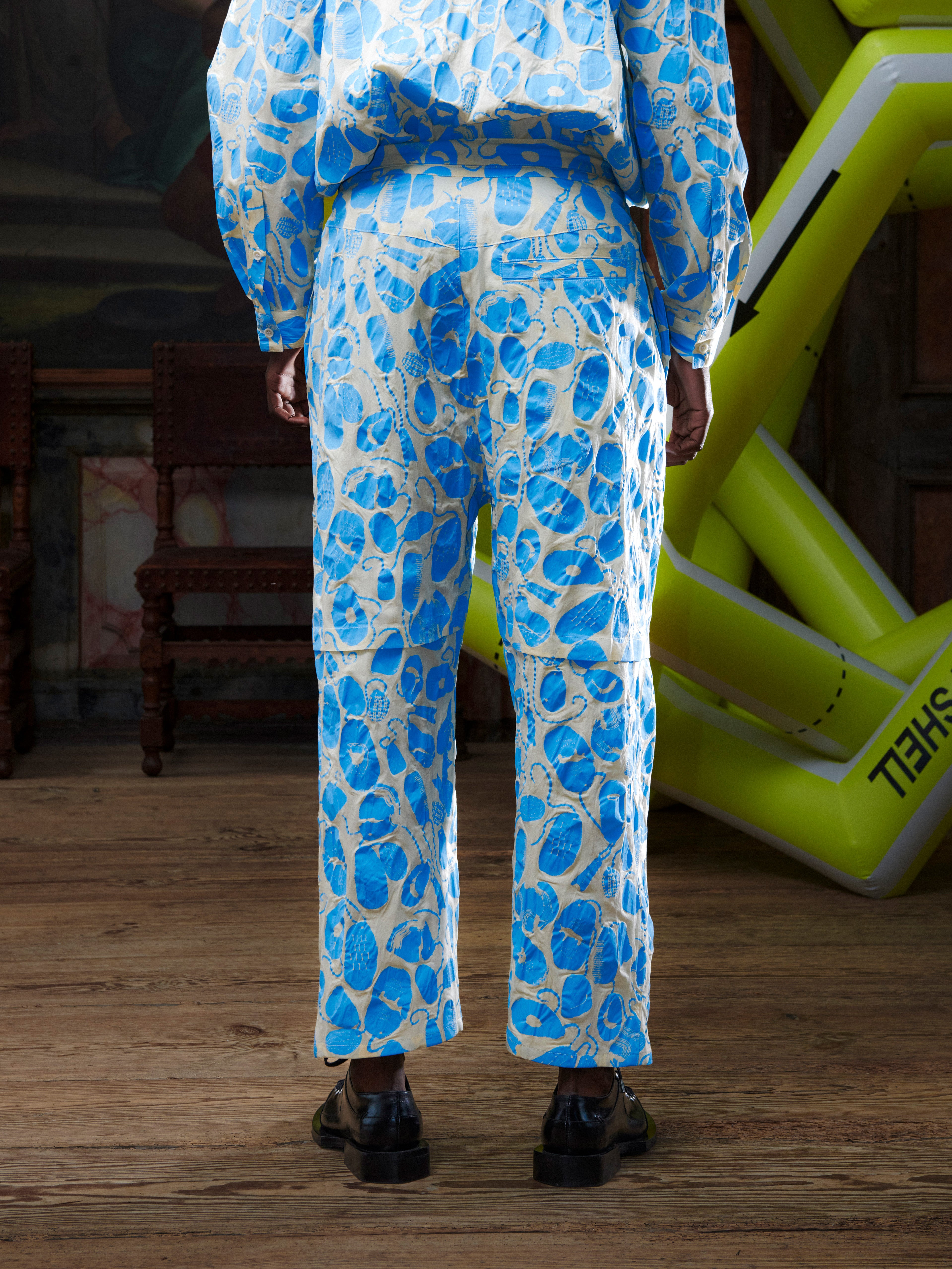 Close-up back view of male model wearing the Drawstring Pants in Ecru Amulets, highlighting the all-over blue amulets pattern and straight-leg back detailing.