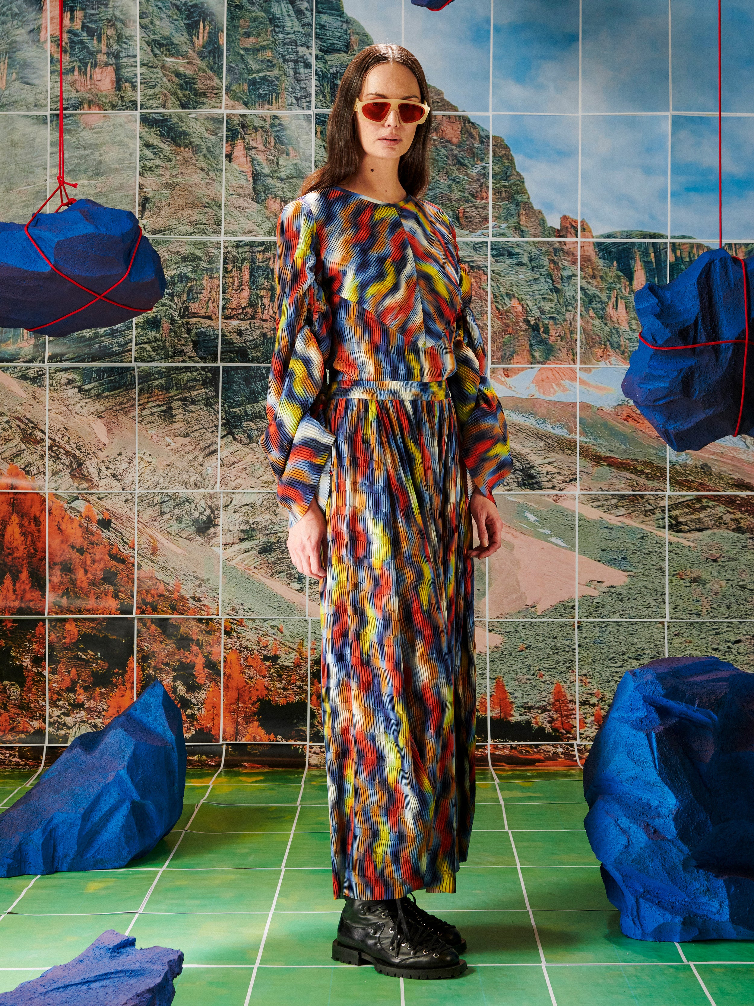 Woman in a colorful pleated top and pants from Henrik Vibskov standing in front of a tiled wall with mountain illustration.