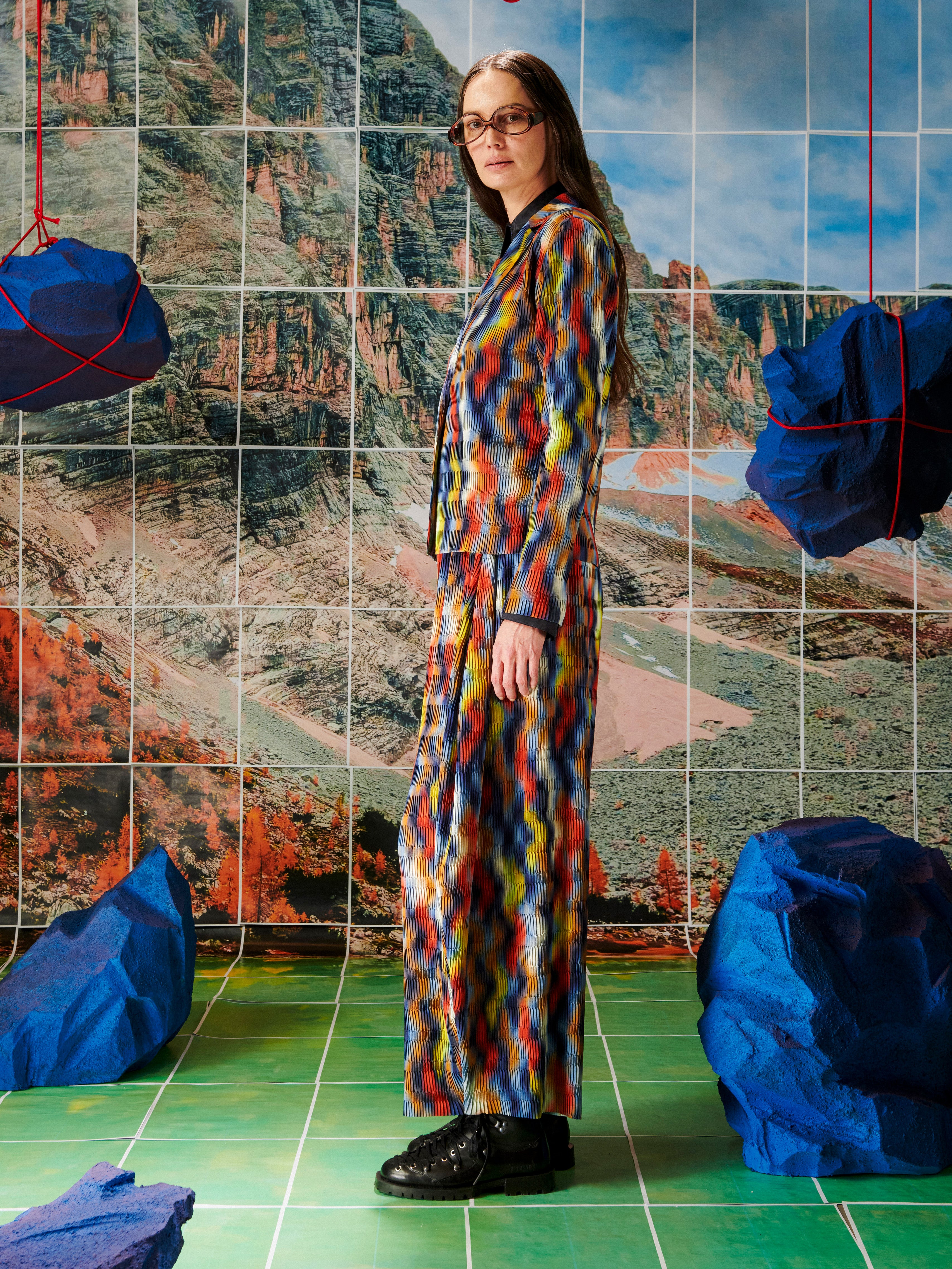 Person wearing a colorful outfit standing in front of a tiled wall with a mountainous landscape.
