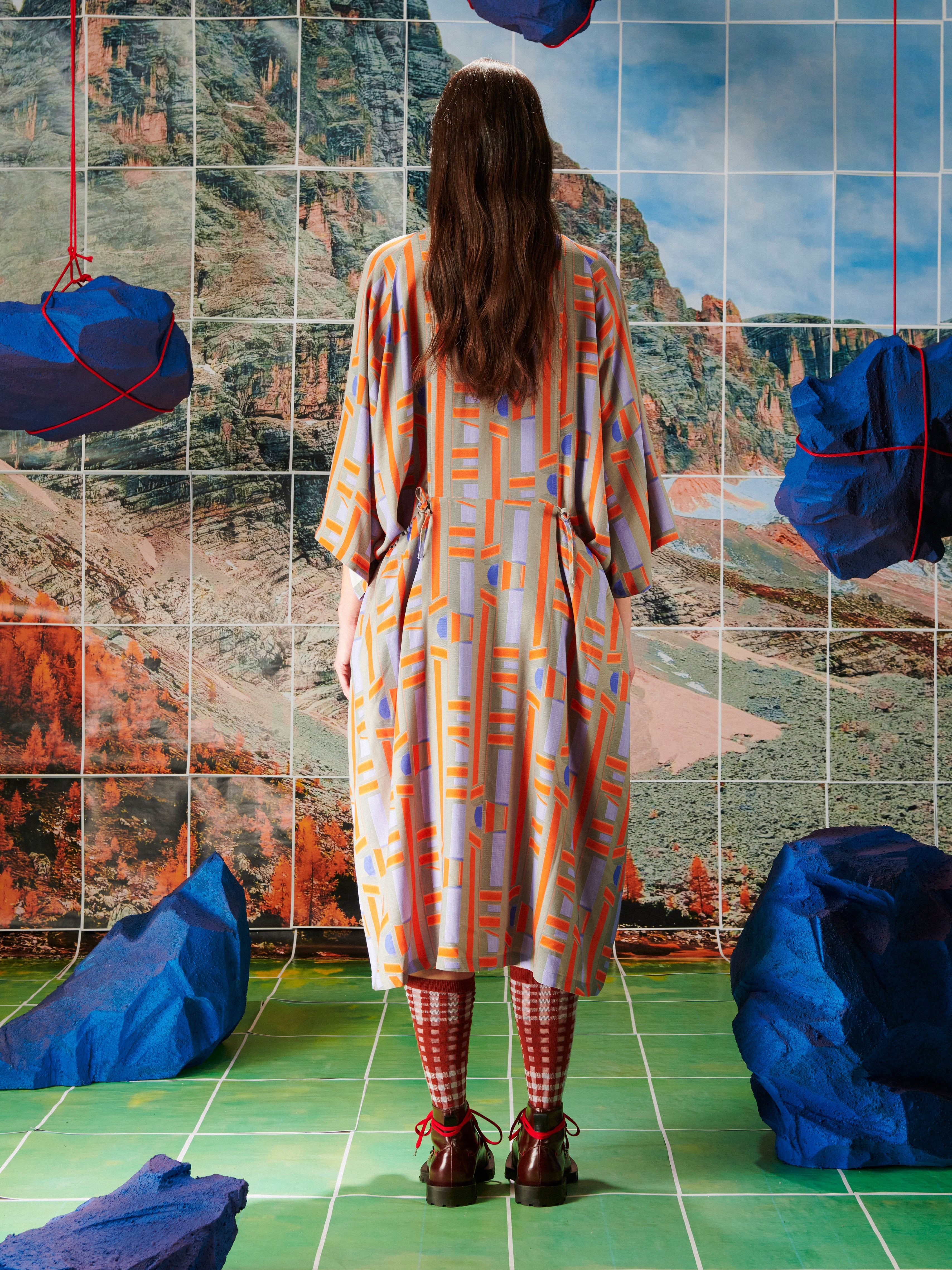 Woman in a patterned dress by Henrik Vibskov standing in front of a tiled wall with abstract designs.