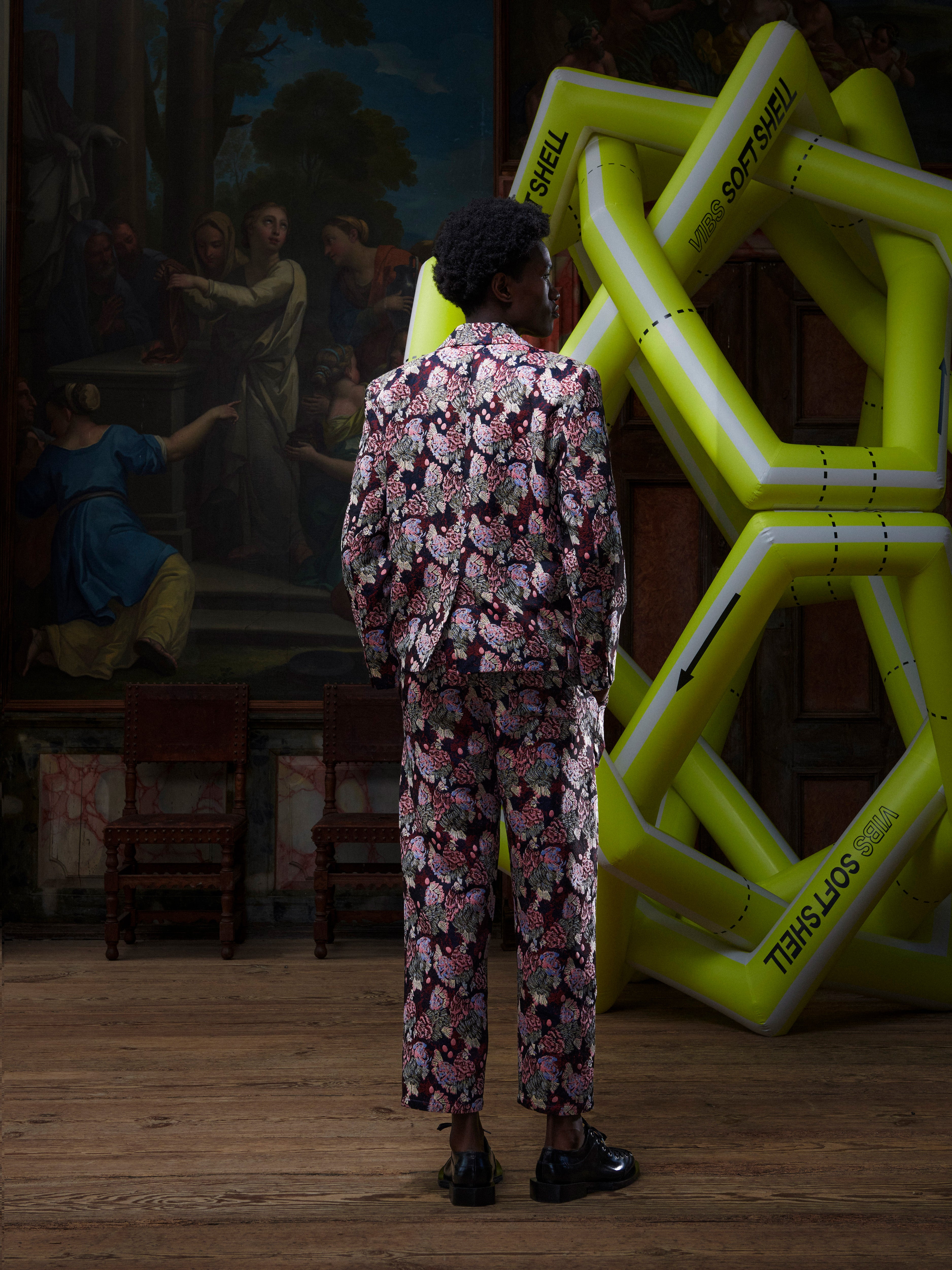 Back view of male model wearing a patterned suit by Henrik Vibskov, standing in front of a large inflatable sculpture with a painting in the background.