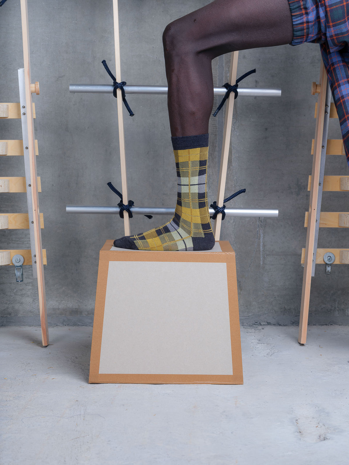 A person's leg wearing a yellow and black plaid sock stands on a wooden platform in front of a gray concrete wall, with wooden and metal display racks in the background.