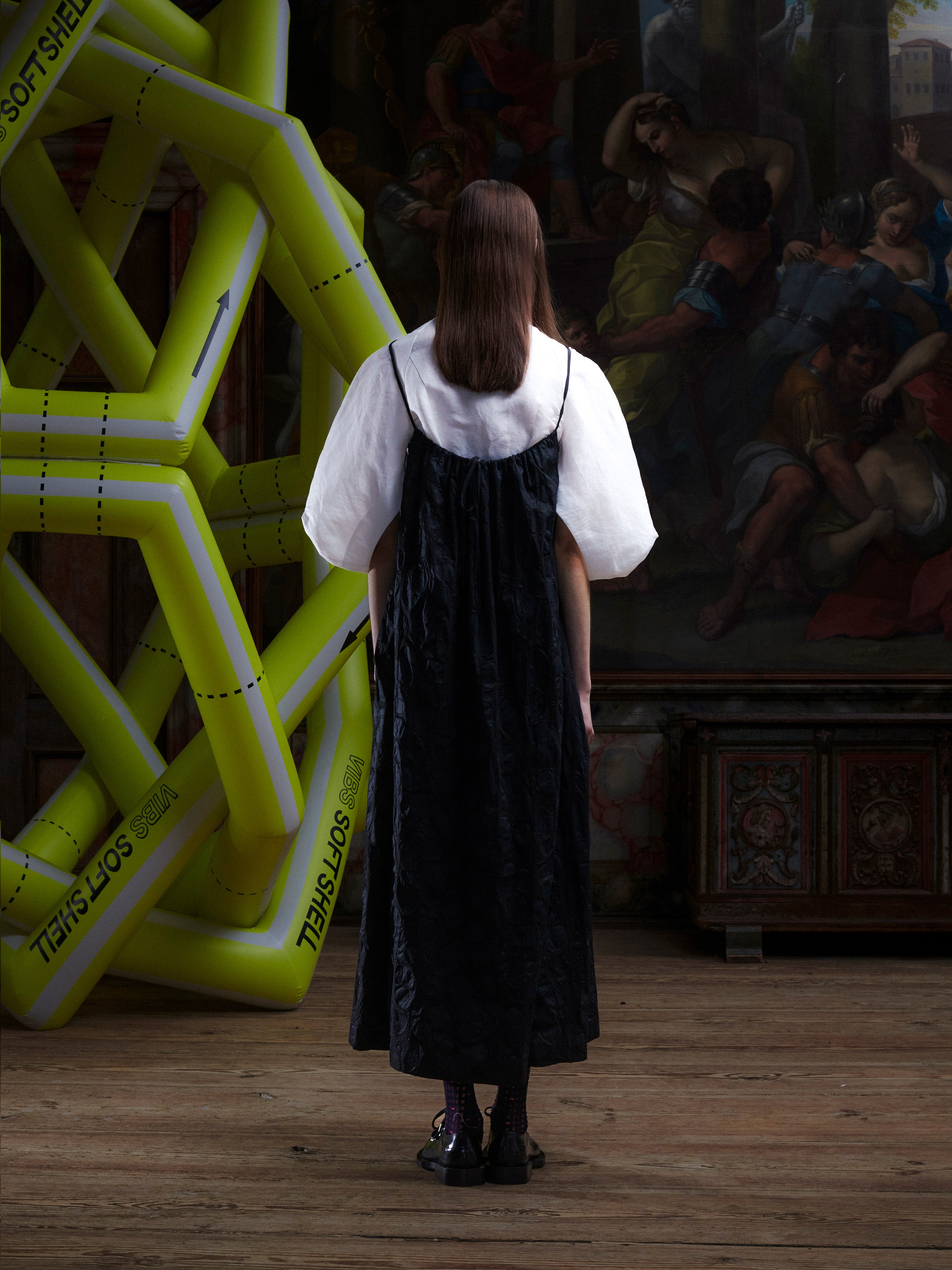 Back view of woman wearing the black-on-black pattern, sleeveless dress on top of a white blouse from Henrik Vibskov.