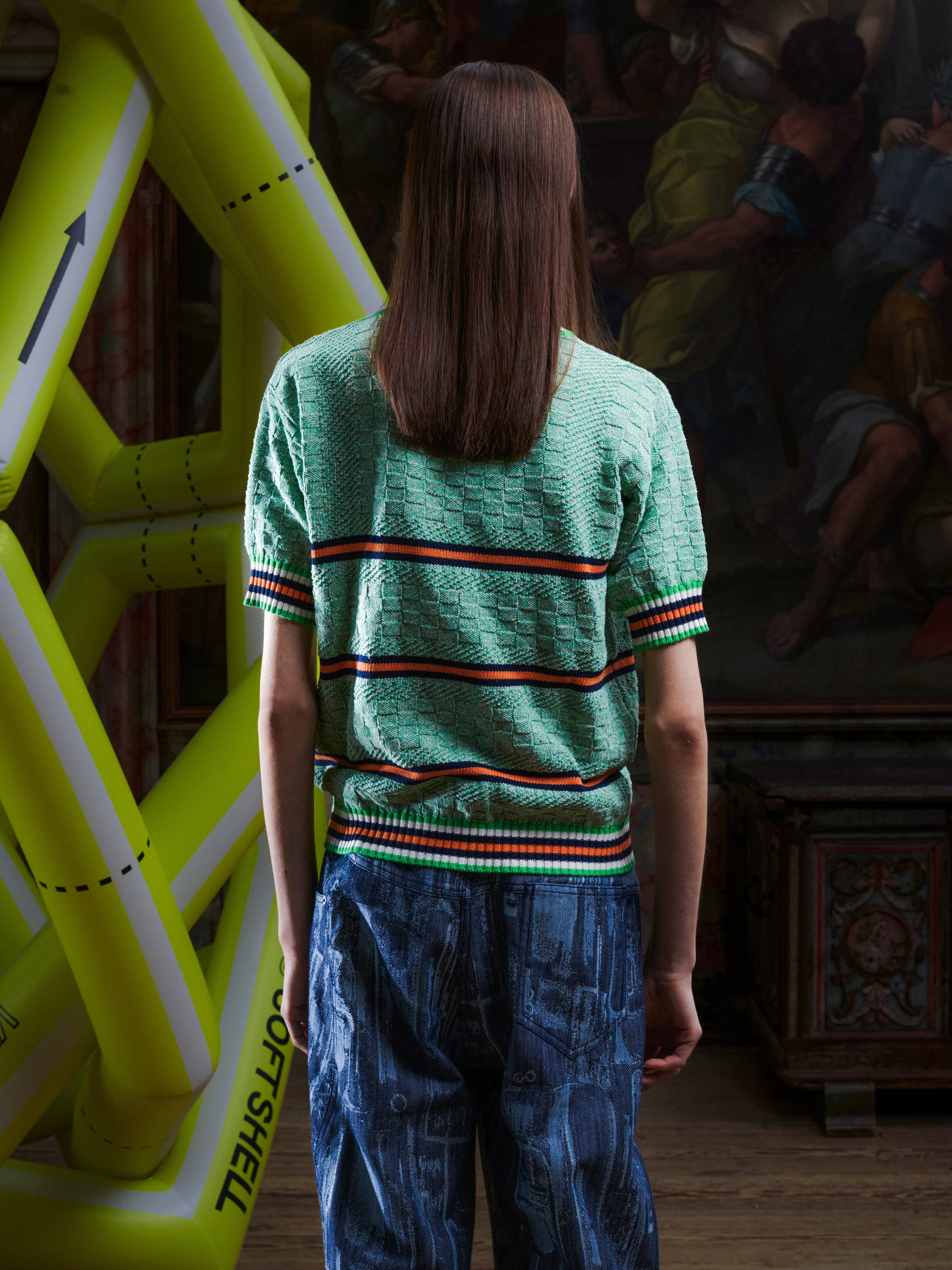 Back view of woman wearing the mint green knitted top with stripe details from Henrik Vibskov with a yellow inflatable sculpture in the background.
