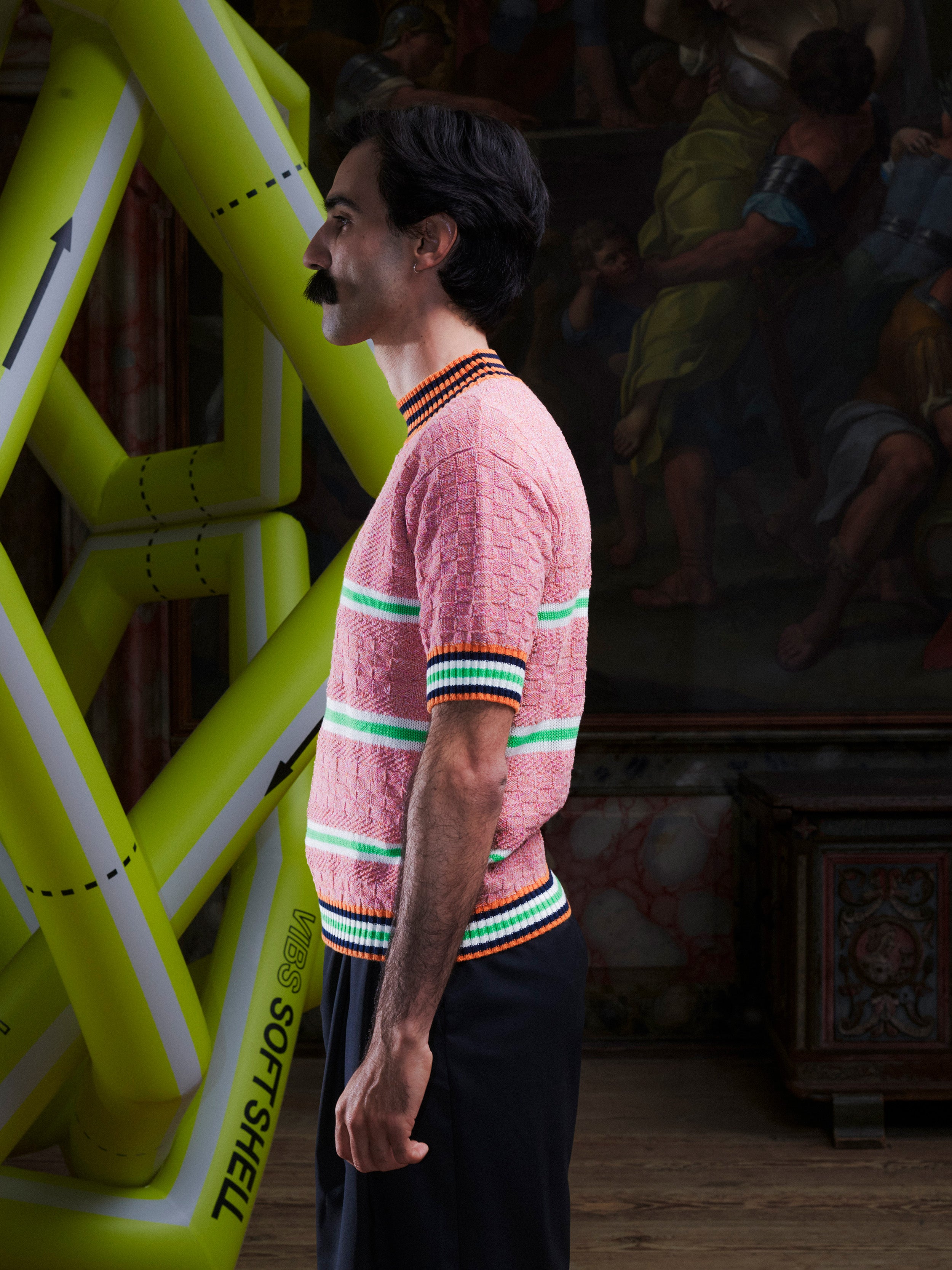 Side profile of man wearing the pink-green knitted top with stripe details from Henrik Vibskov with a yellow inflatable sculpture in the background.