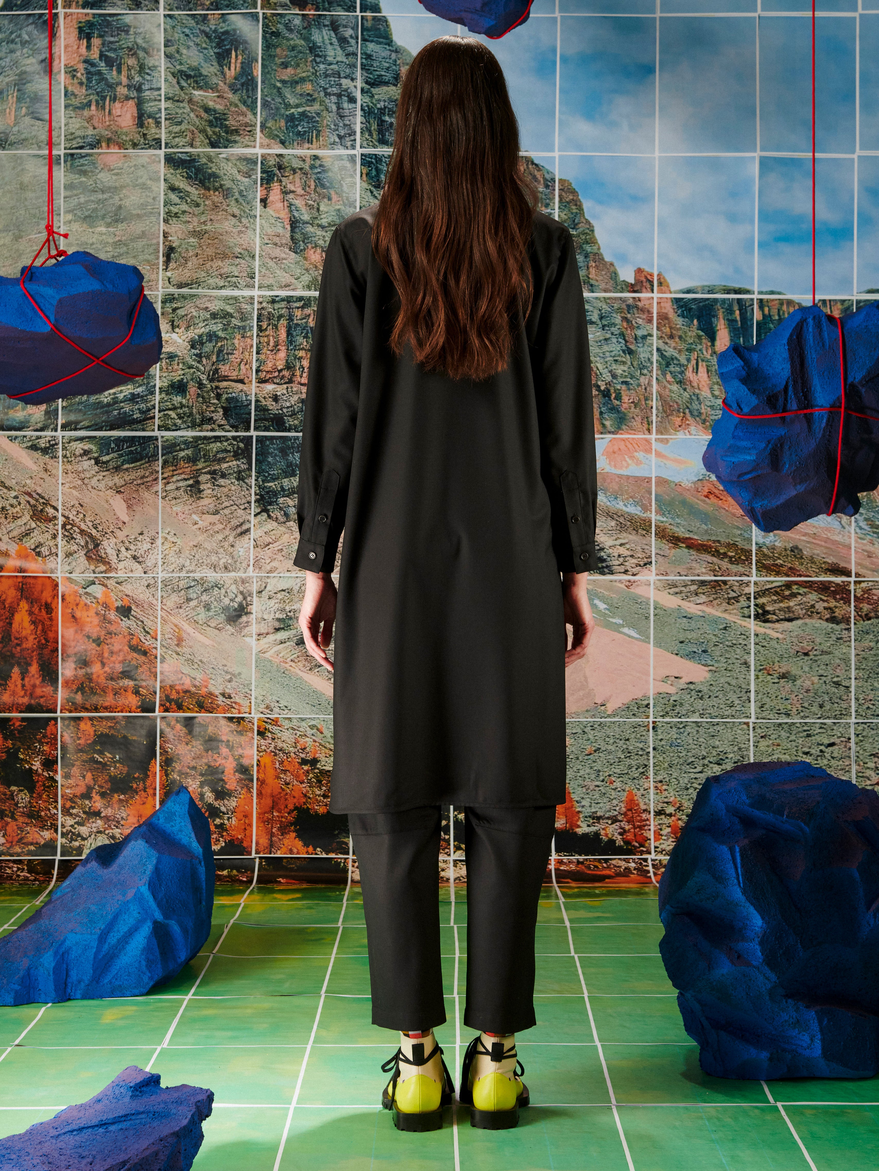 Person wearing a dark outfit standing in front of a tiled wall with abstract patterns and blue sculptures.