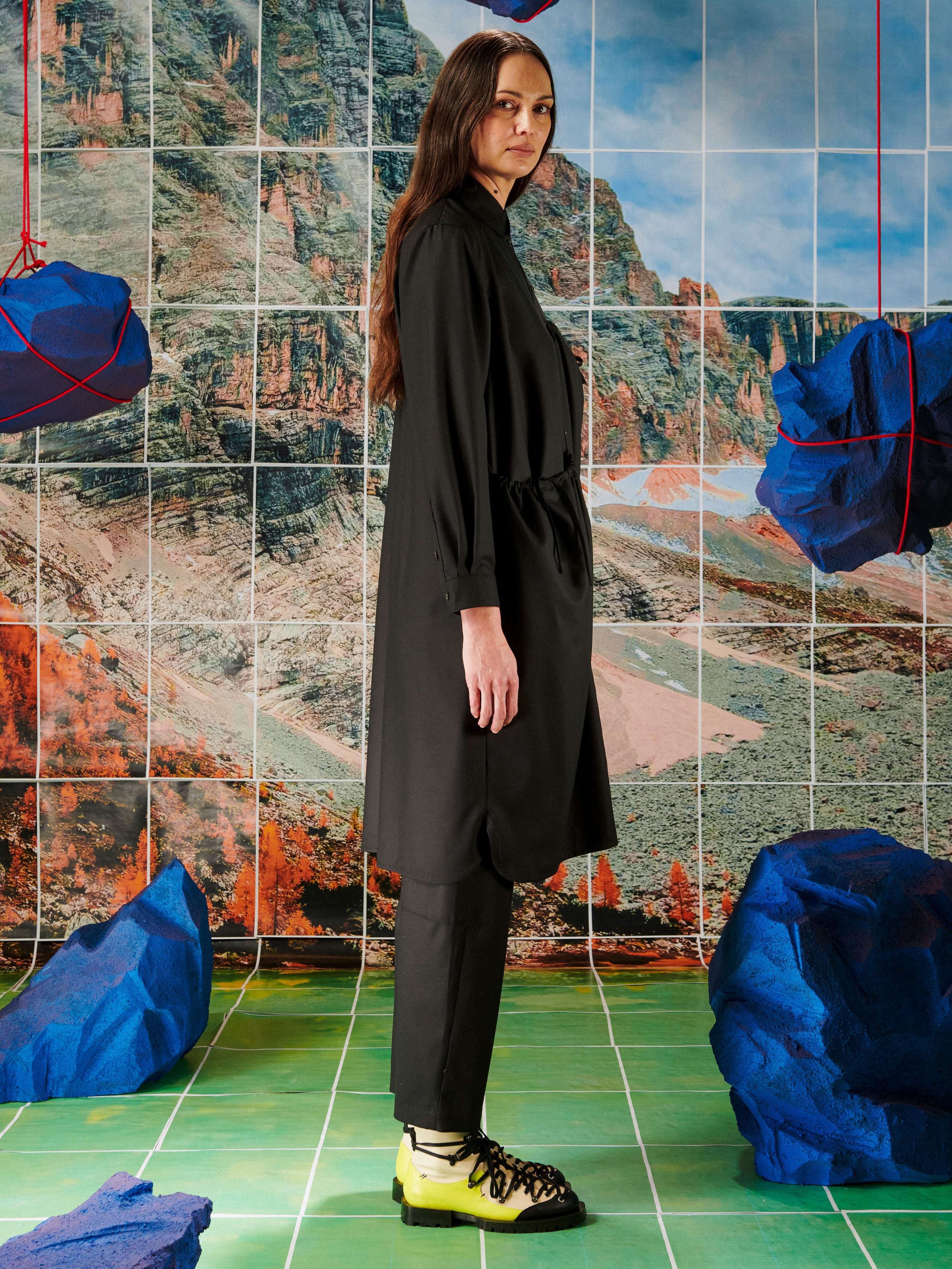 Person wearing a black outfit standing in front of a tiled wall with a scenic mosaic.