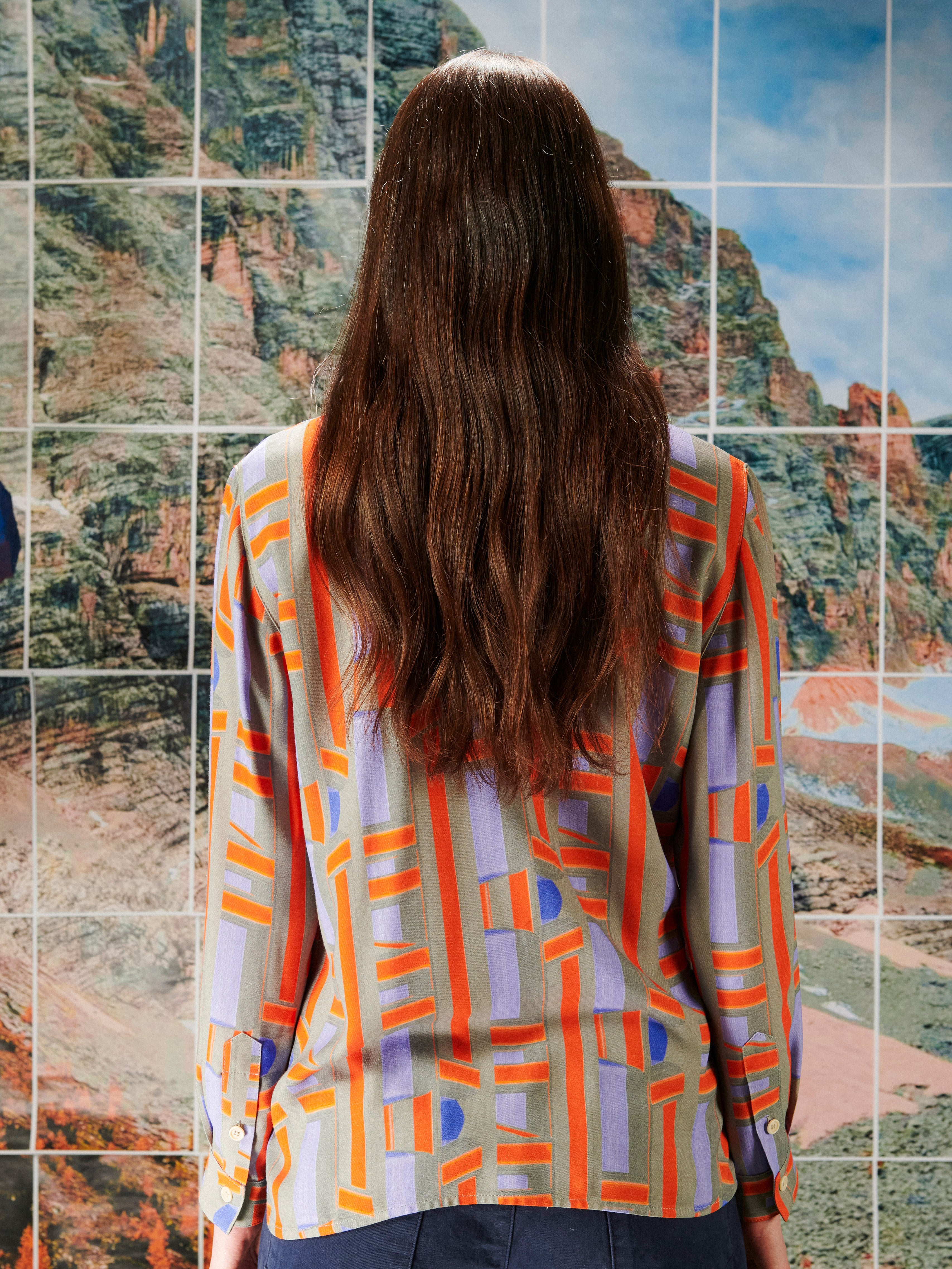 Person wearing a colorful patterned shirt from Henrik Vibskov standing in front of a mosaic of landscape images.