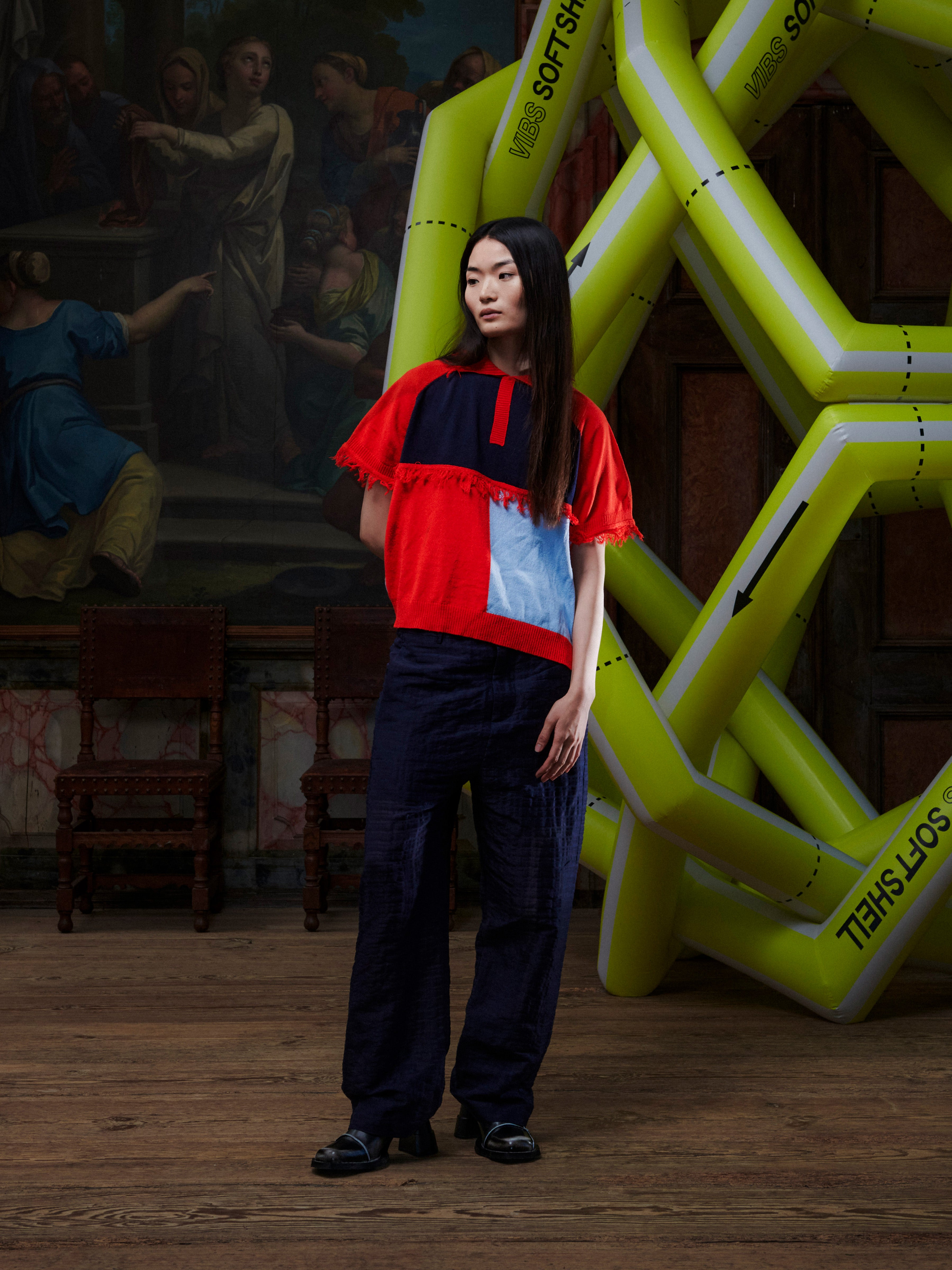 Person wearing a bright red knitted short sleeve shirt standing in front of a large inflatable structure and classical painting.