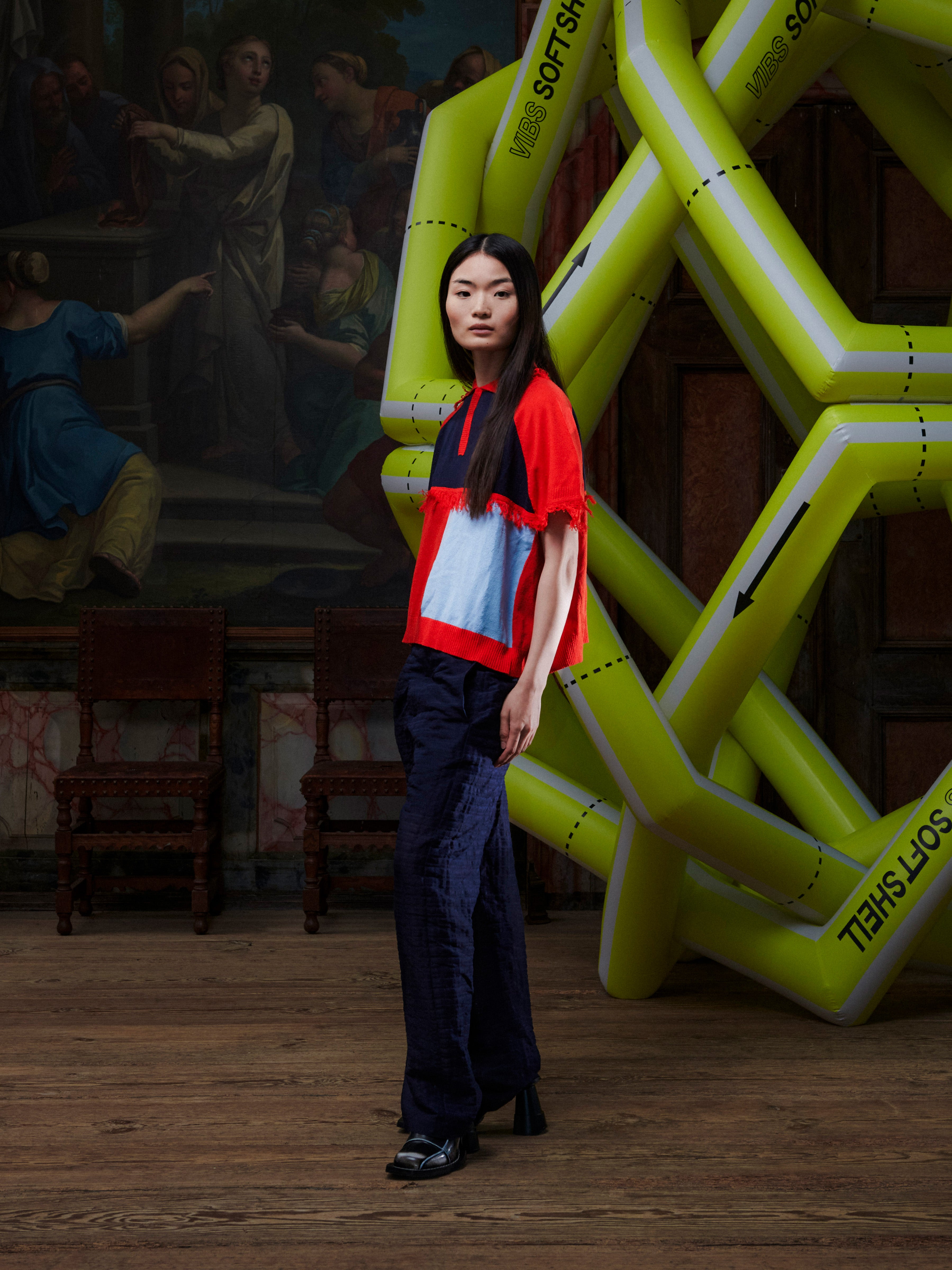 Side profile of person wearing a bright red knitted short sleeve shirt standing in front of a large inflatable structure and classical painting.