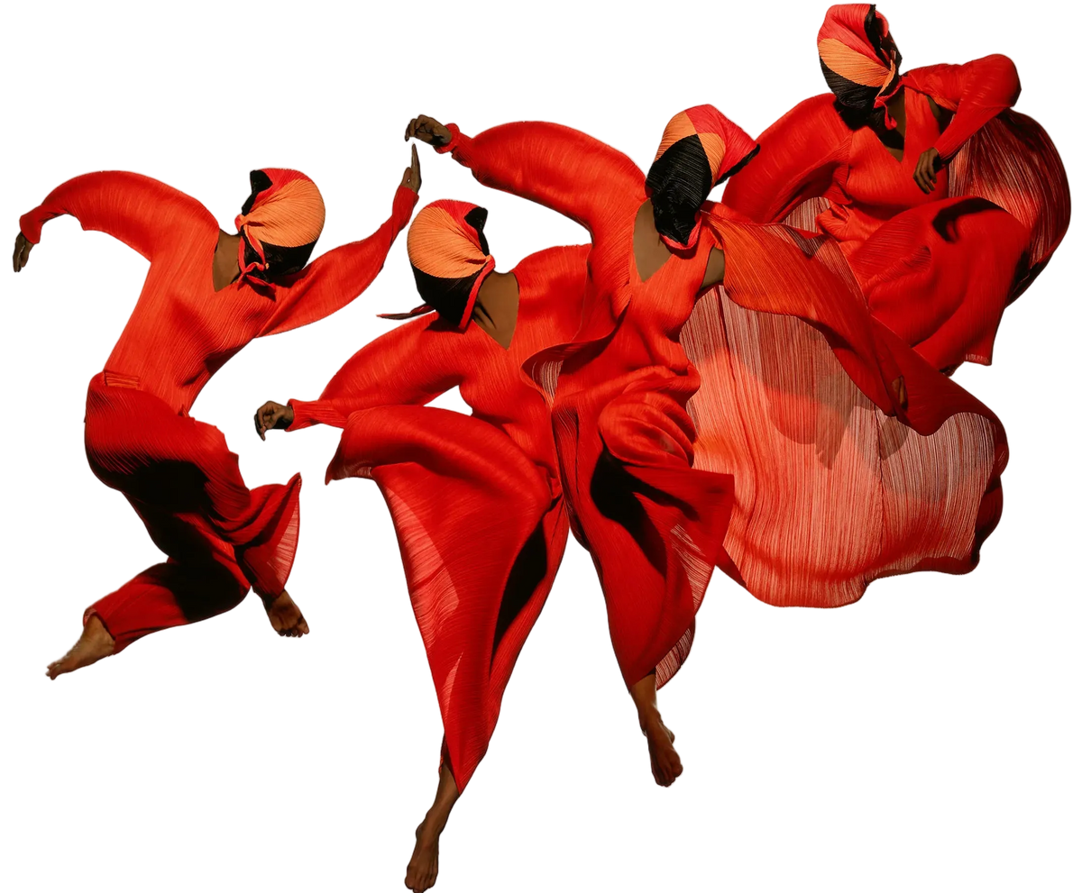 Group of dancers in bright red pleated pleats garments and sculptural headpieces, captured mid-movement against a light background.