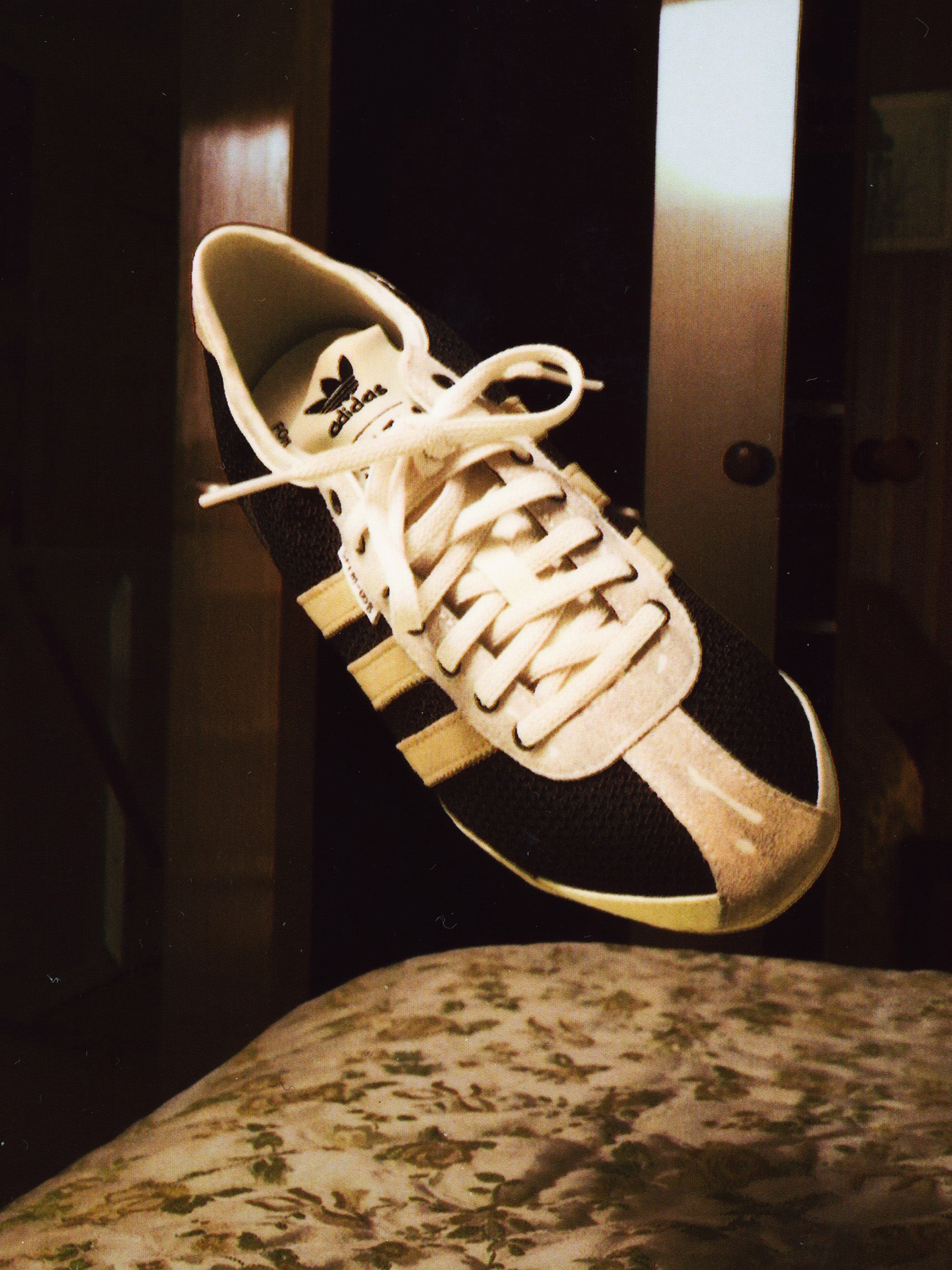Adidas Tokyo W SFTM in Black/Beige/Gold - campaign shot showing sneaker suspended in mid-air with beige three stripes and off-white lace-up detail against a dark interior background