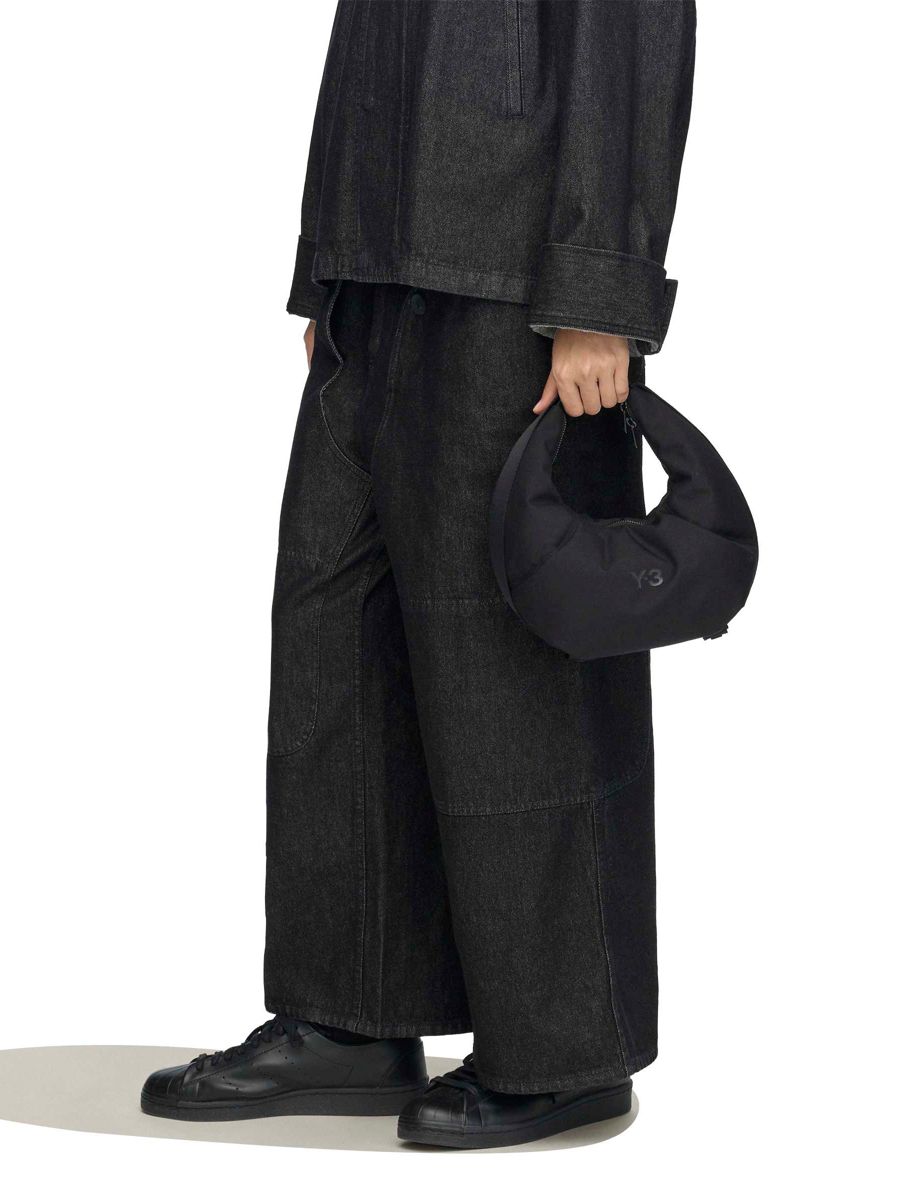 Y-3 Crescent Bag on-model close-up, hand carry grip, crescent silhouette at hip, black wide-leg trousers styling context, adjustable webbing strap