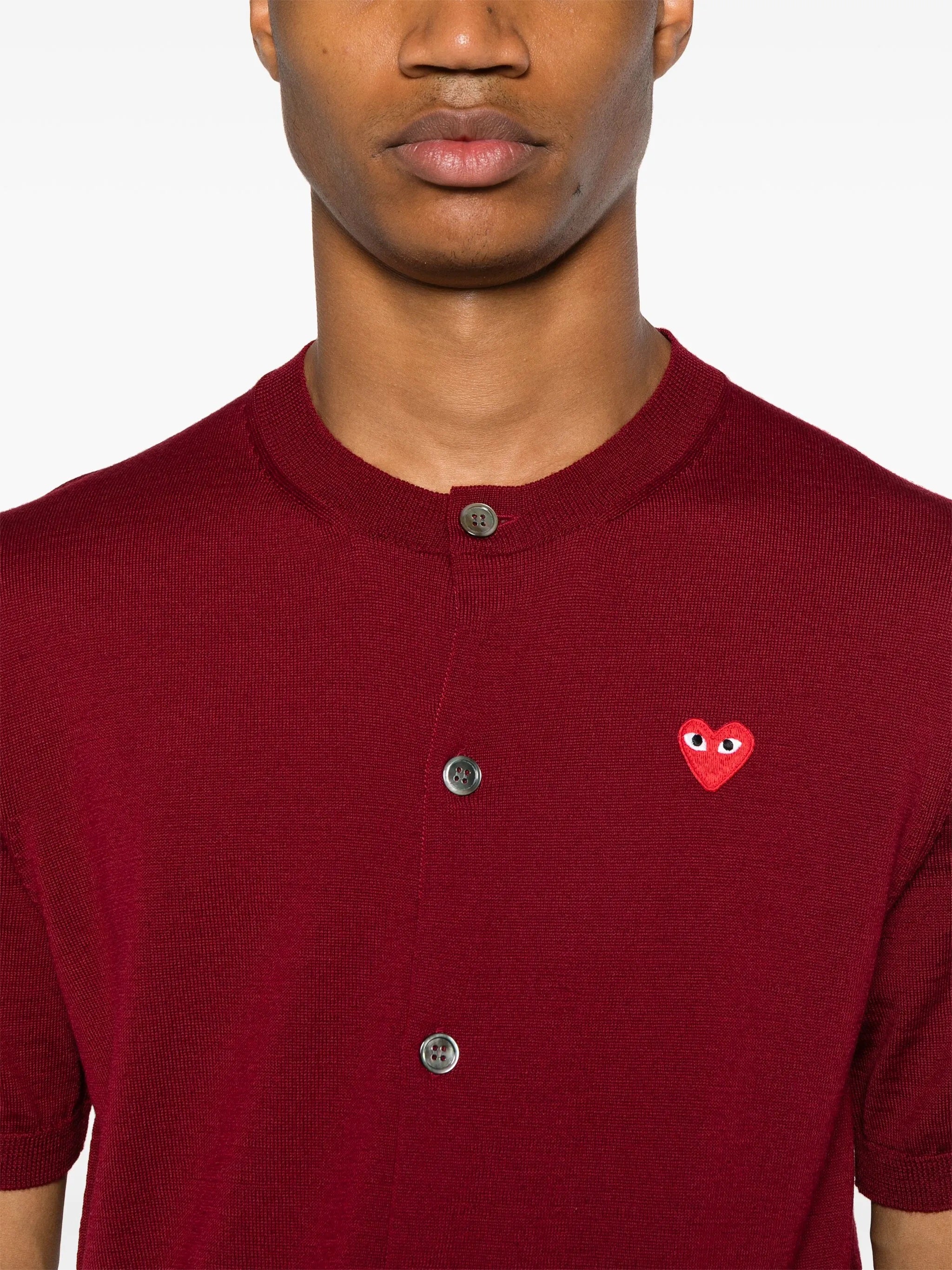 Close-up of person wearing the red short-sleeve cardigan with a red heart logo from Comme des Garcons Play on a white background