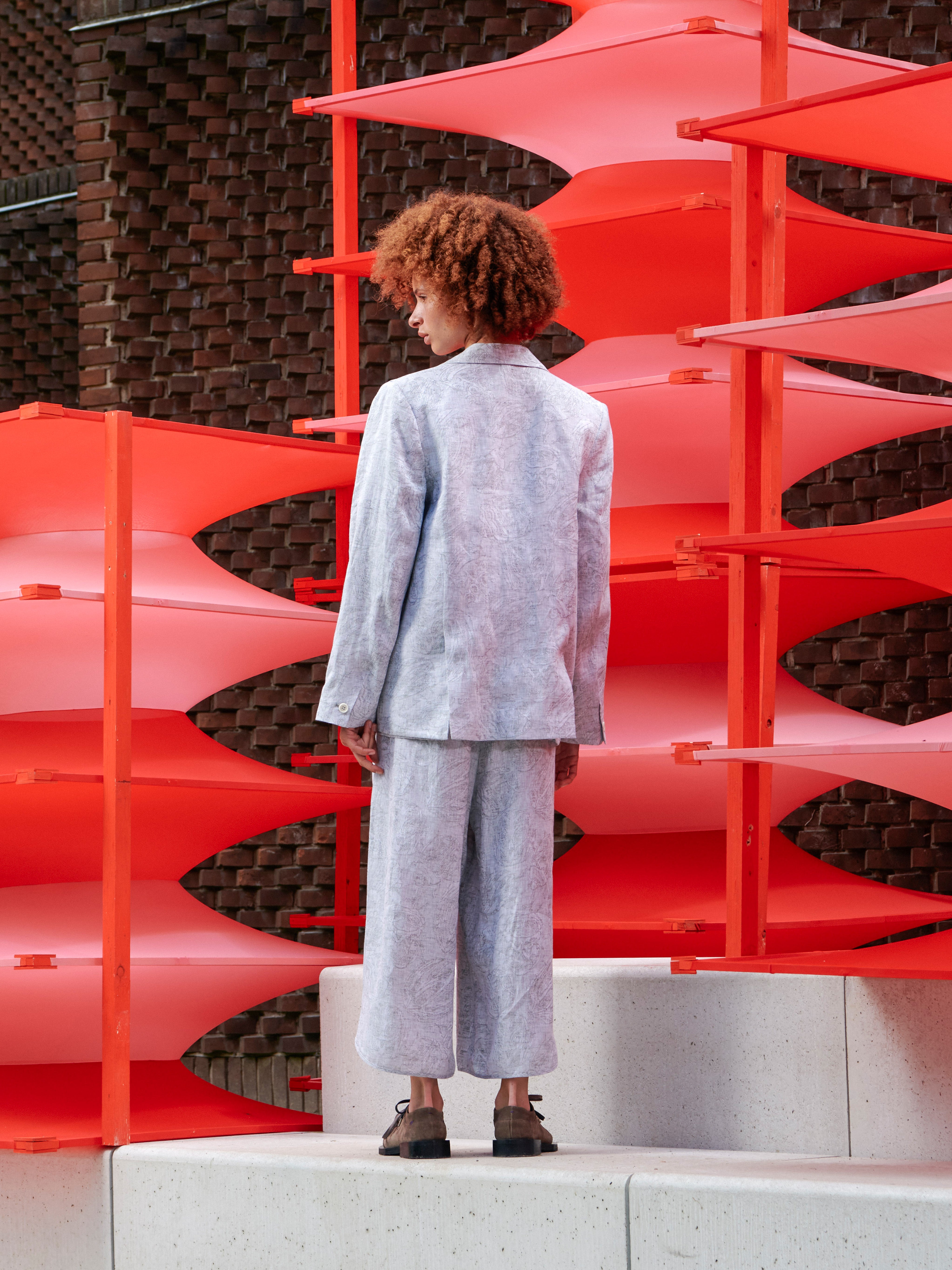 Back view of a model wearing the blazer, showing the drape, layered construction, and grey floral textured pattern.