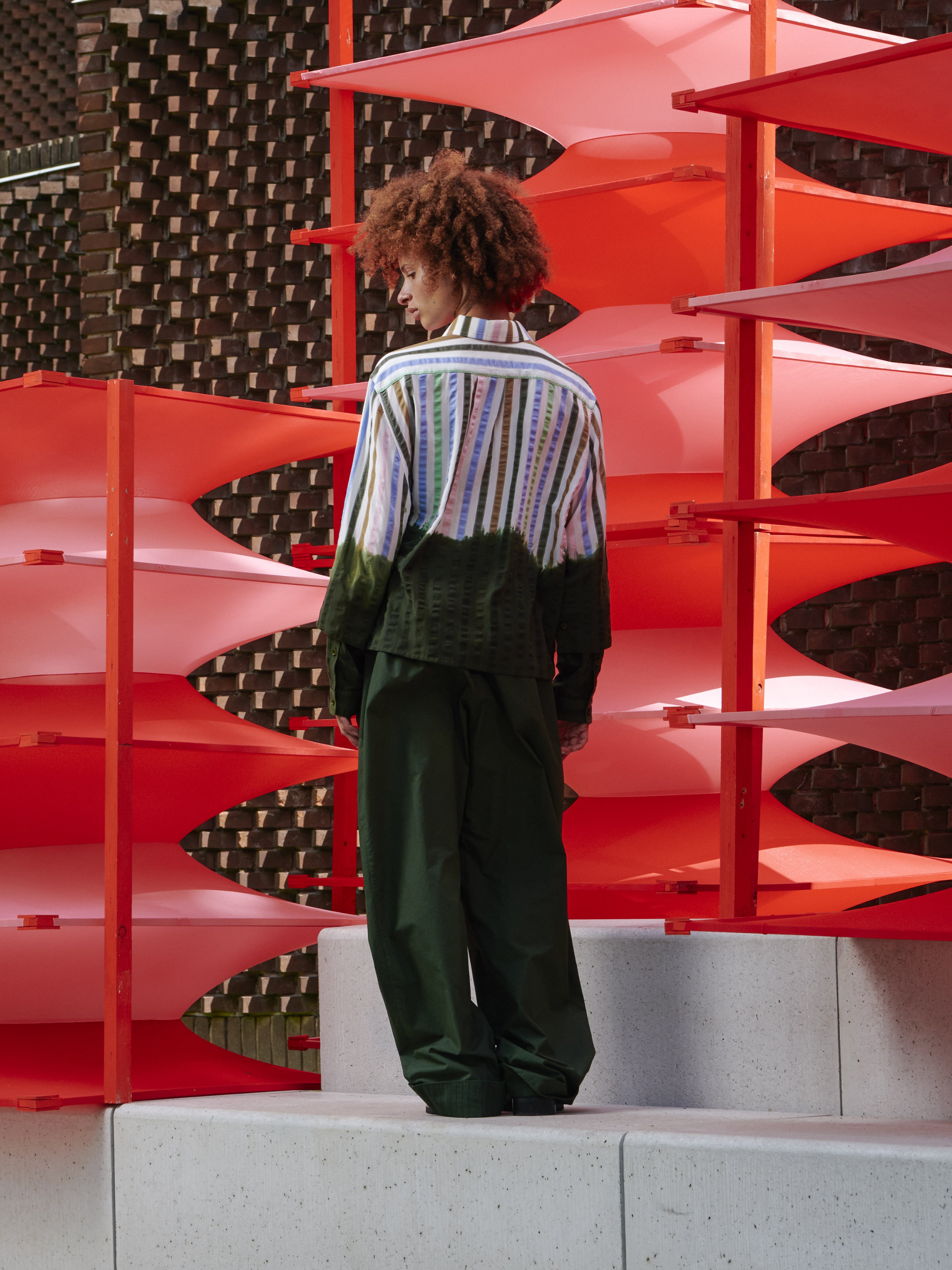 Model wearing Blousse Shirt in DipDye Stripes by Henrik Vibskov, back view showing full-length multicolour stripe print with deep forest green dip-dye hem, relaxed fit and long sleeves, Spring/Summer 2026 Key Collection