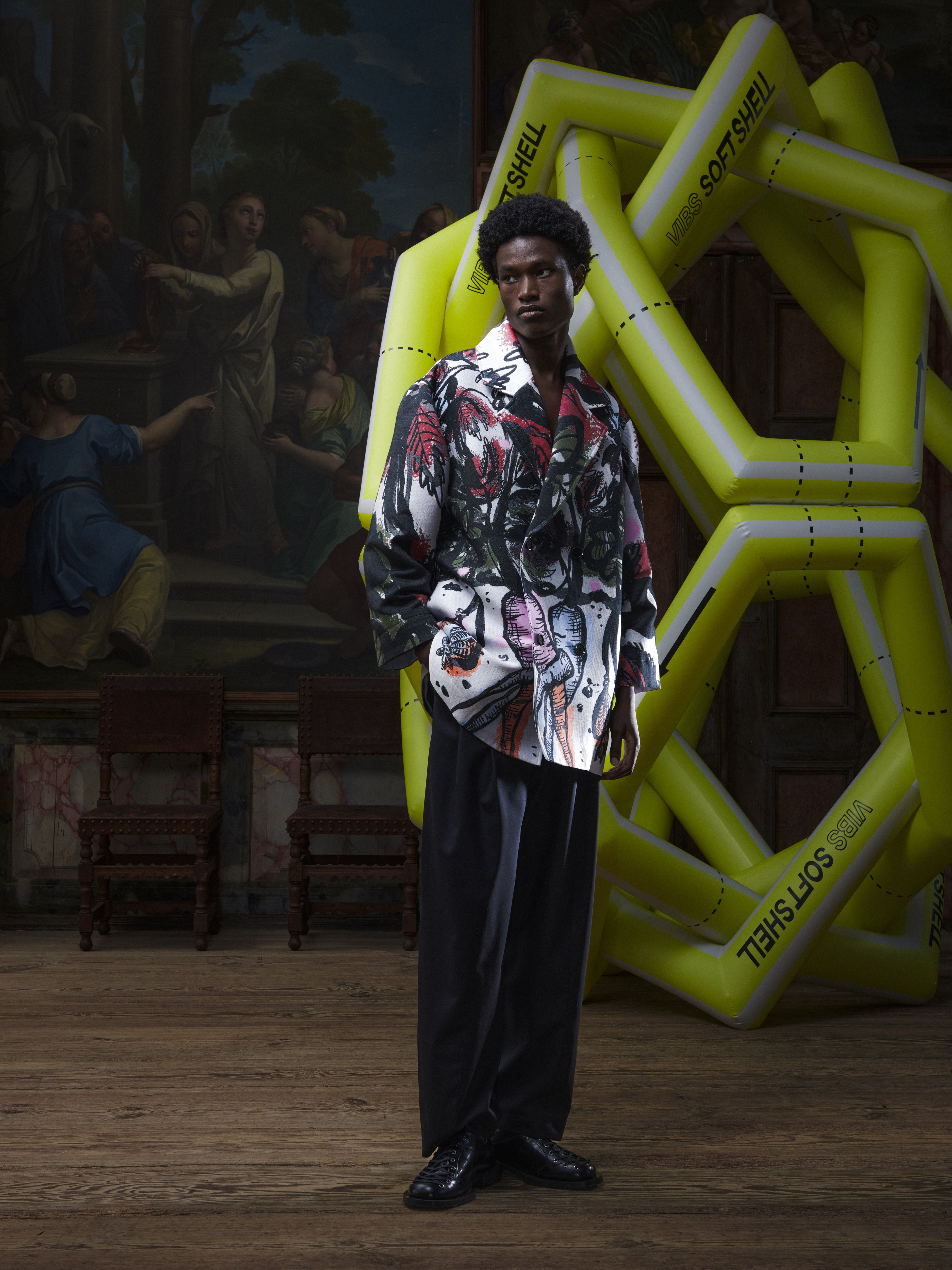 Front view of the male model wearing the Care Coat in Carrots from Henrik Vibskov Spring/Summer 2026 Studio Collection, displaying the multicoloured carrot print, double-breasted front, long sleeves, and pockets.