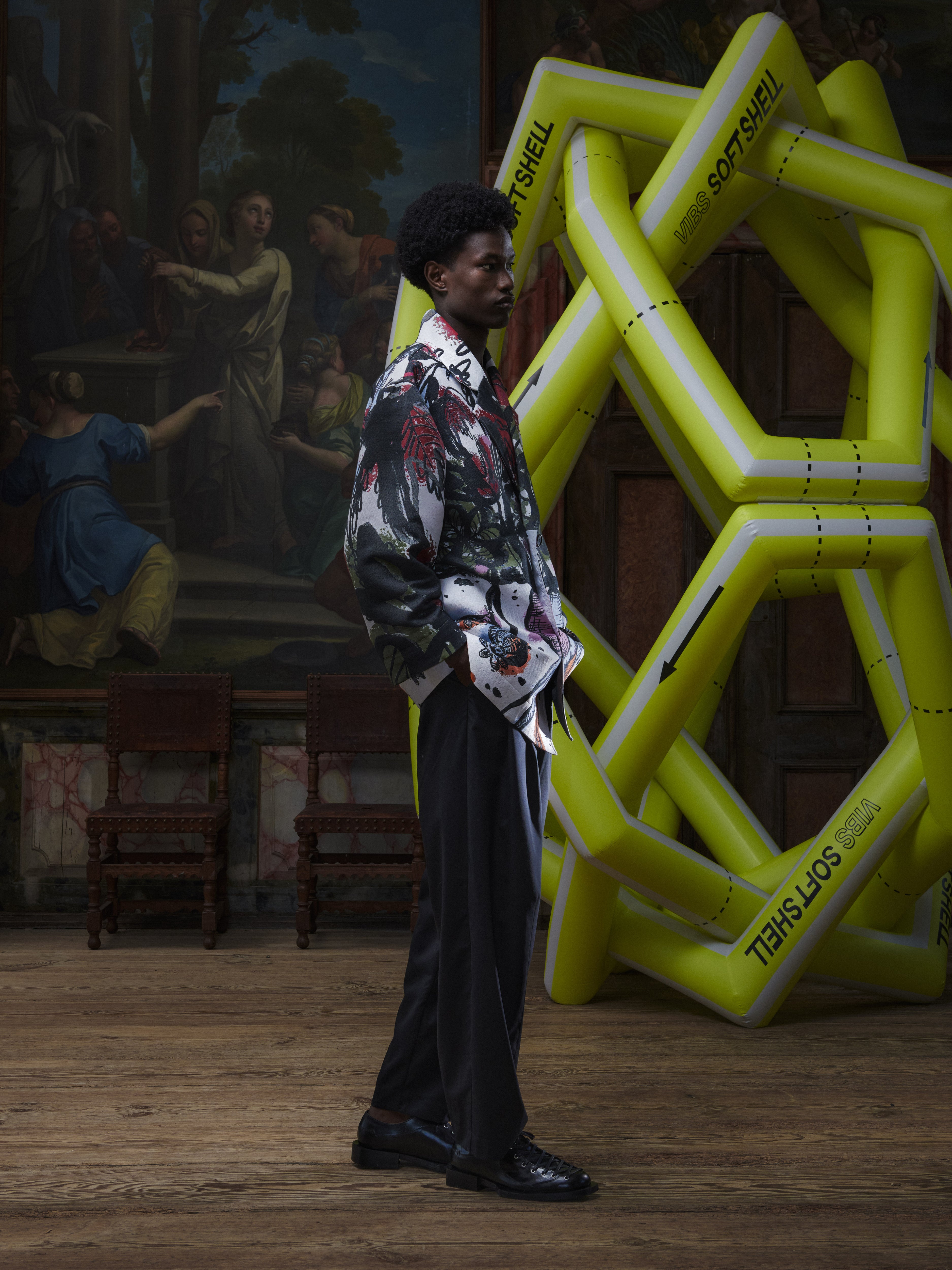 Side view of the male model wearing the Care Coat in Carrots from Henrik Vibskov Spring/Summer 2026 Studio Collection, showing the colorful carrot pattern, long-sleeve shape, and coat length.