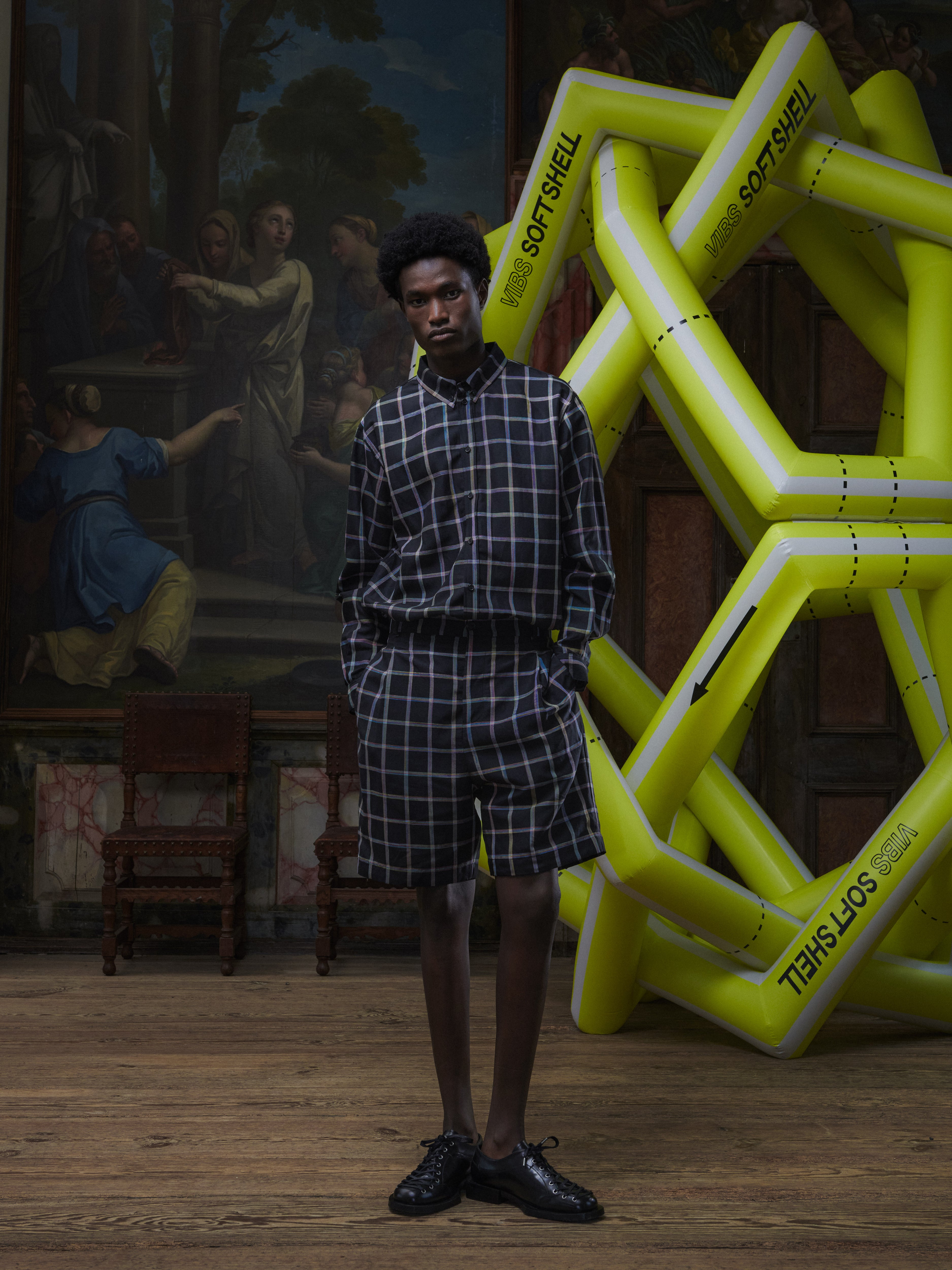Front full-body view of a male model wearing the Claus Shorts in Black Checks from Henrik Vibskov Spring/Summer 2026 Studio Collection, highlighting the multicoloured check pattern, side pockets, and relaxed unisex fit.