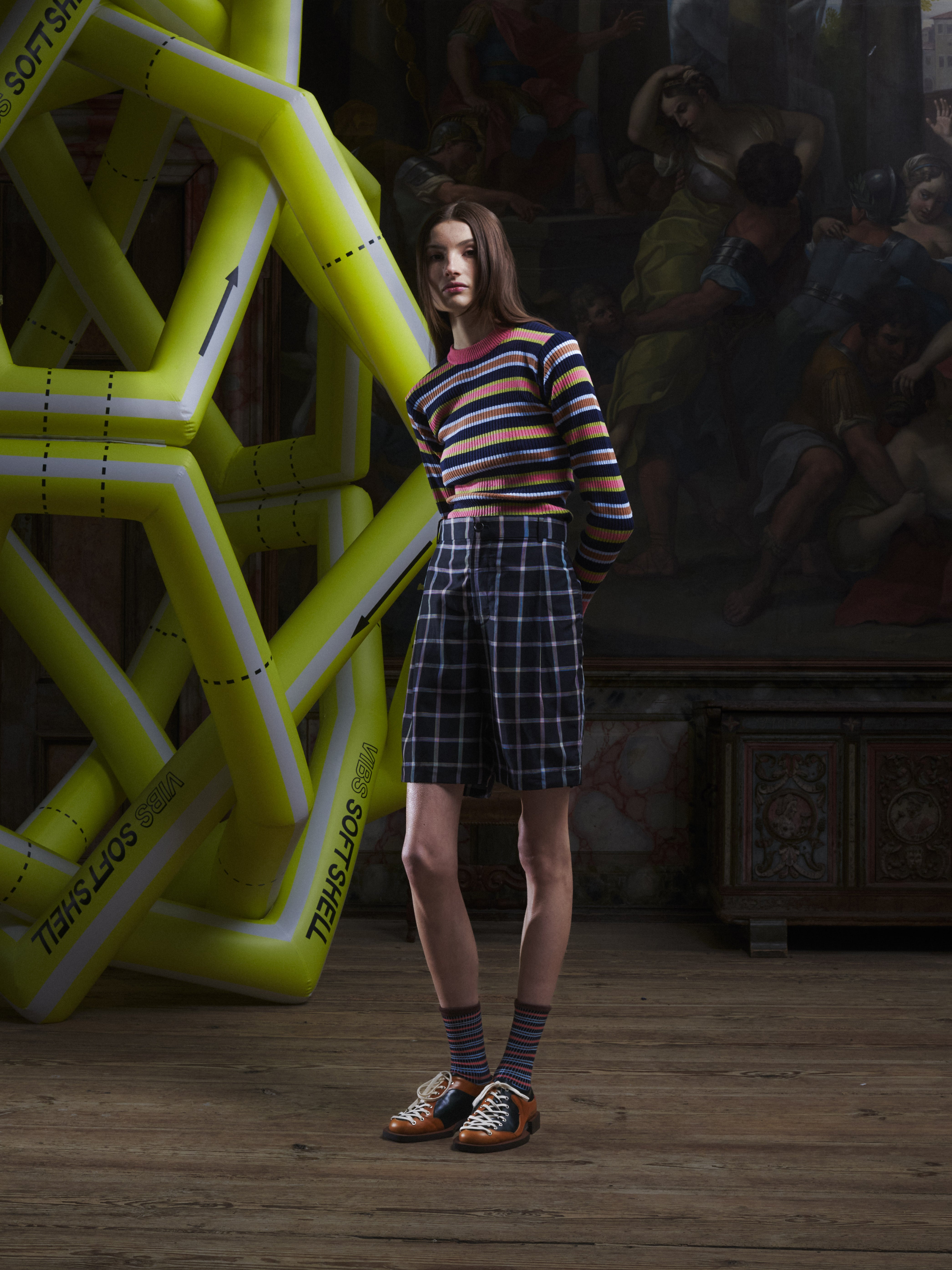 Front view of a female model wearing the Claus Shorts in Black Checks from Henrik Vibskov Spring/Summer 2026 Studio Collection, showing the multicoloured check pattern, front closure, and unisex fit.