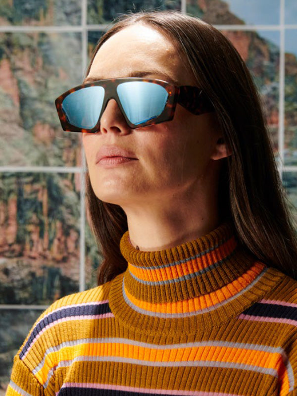 Person wearing reflective sunglasses and a striped sweater in front of a scenic background