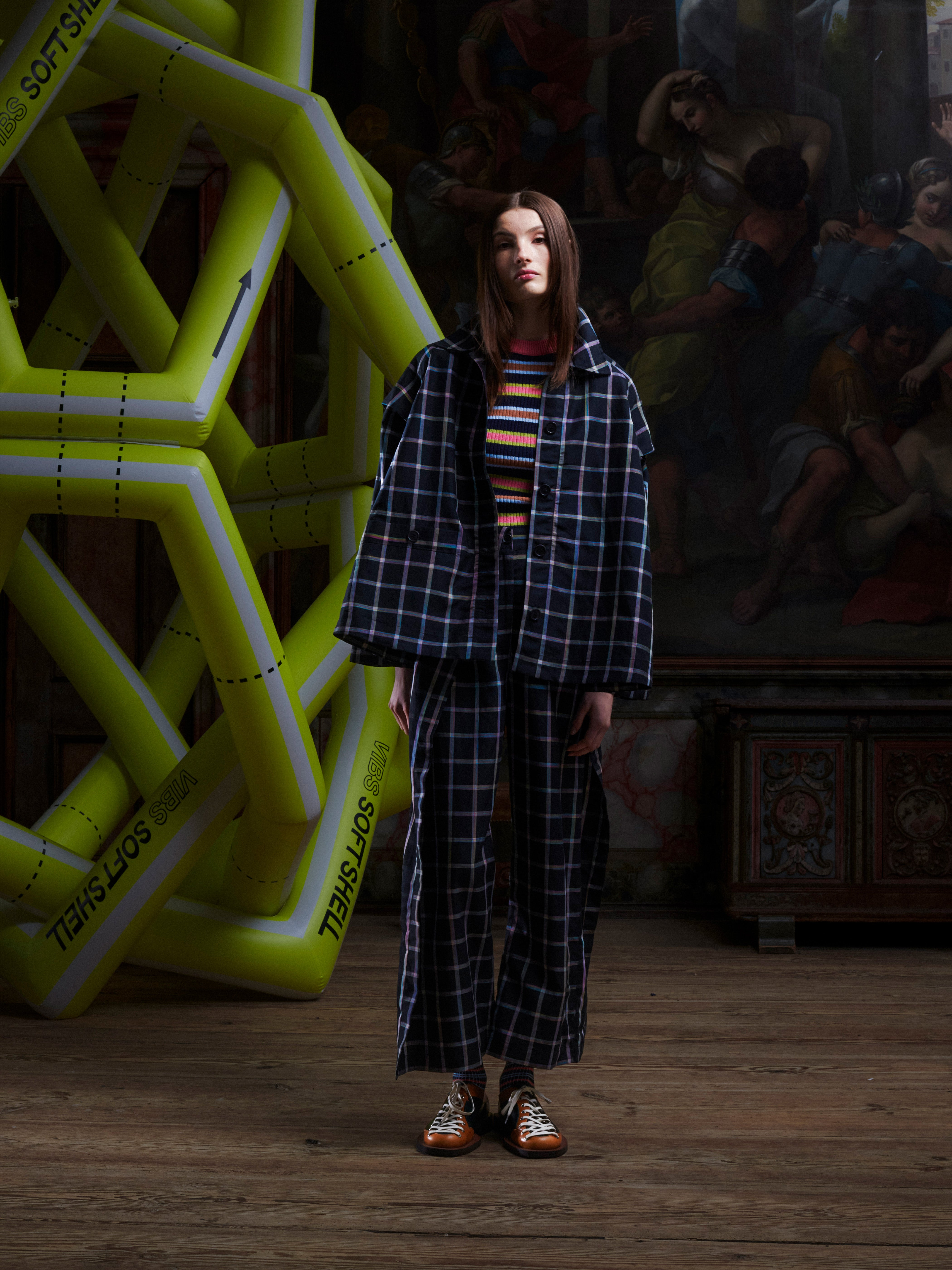 Front view of a female model wearing the Cover Coat in Black Checks from Henrik Vibskov Spring/Summer 2026 Studio Collection, showing the collar, front button closure, and check pattern in a square fit.