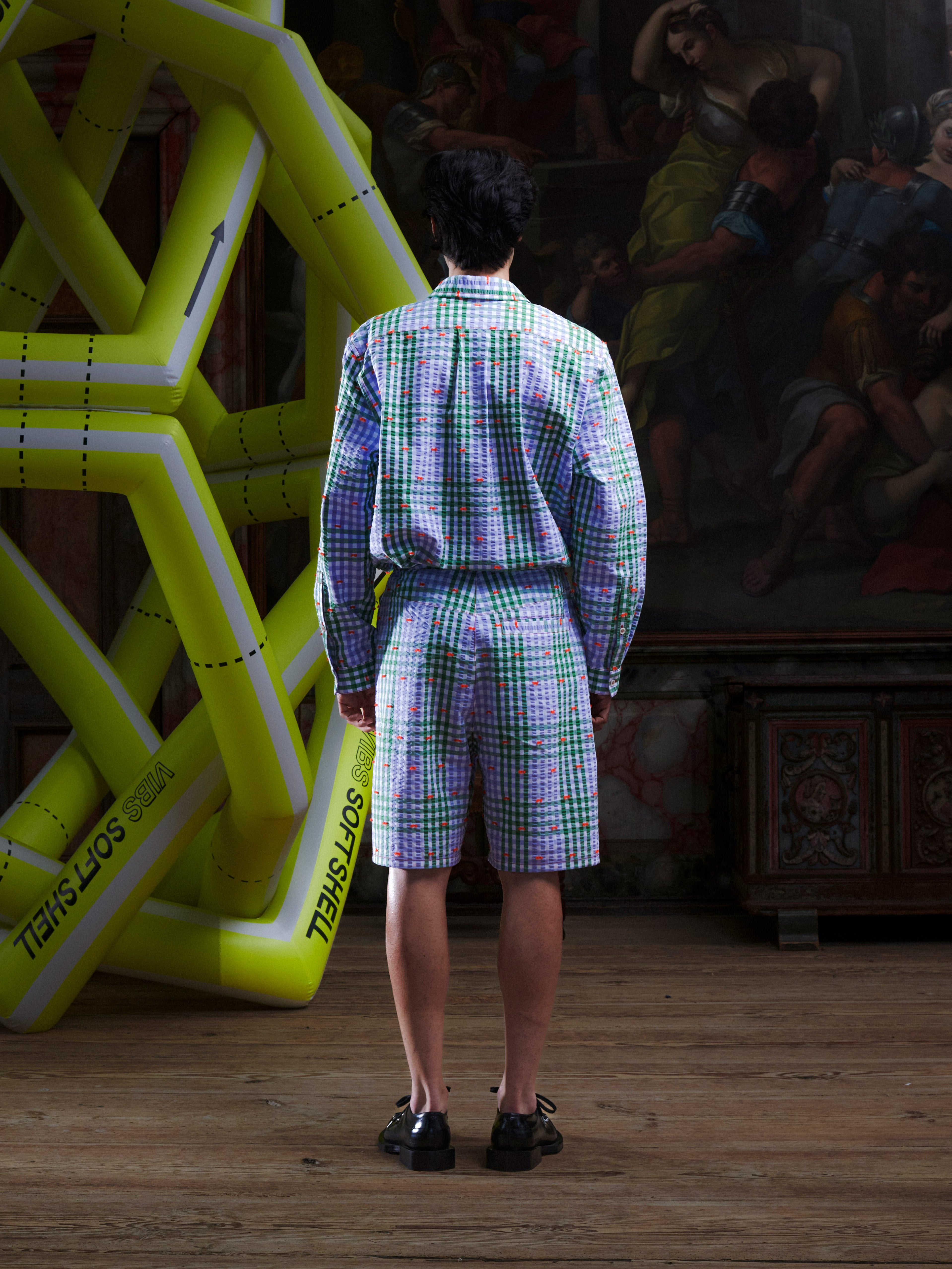 Male model wearing the Drawstring Shorts in Checks Fillé, back view, showing the purple and green checked pattern, knee-length silhouette, and relaxed fit.
