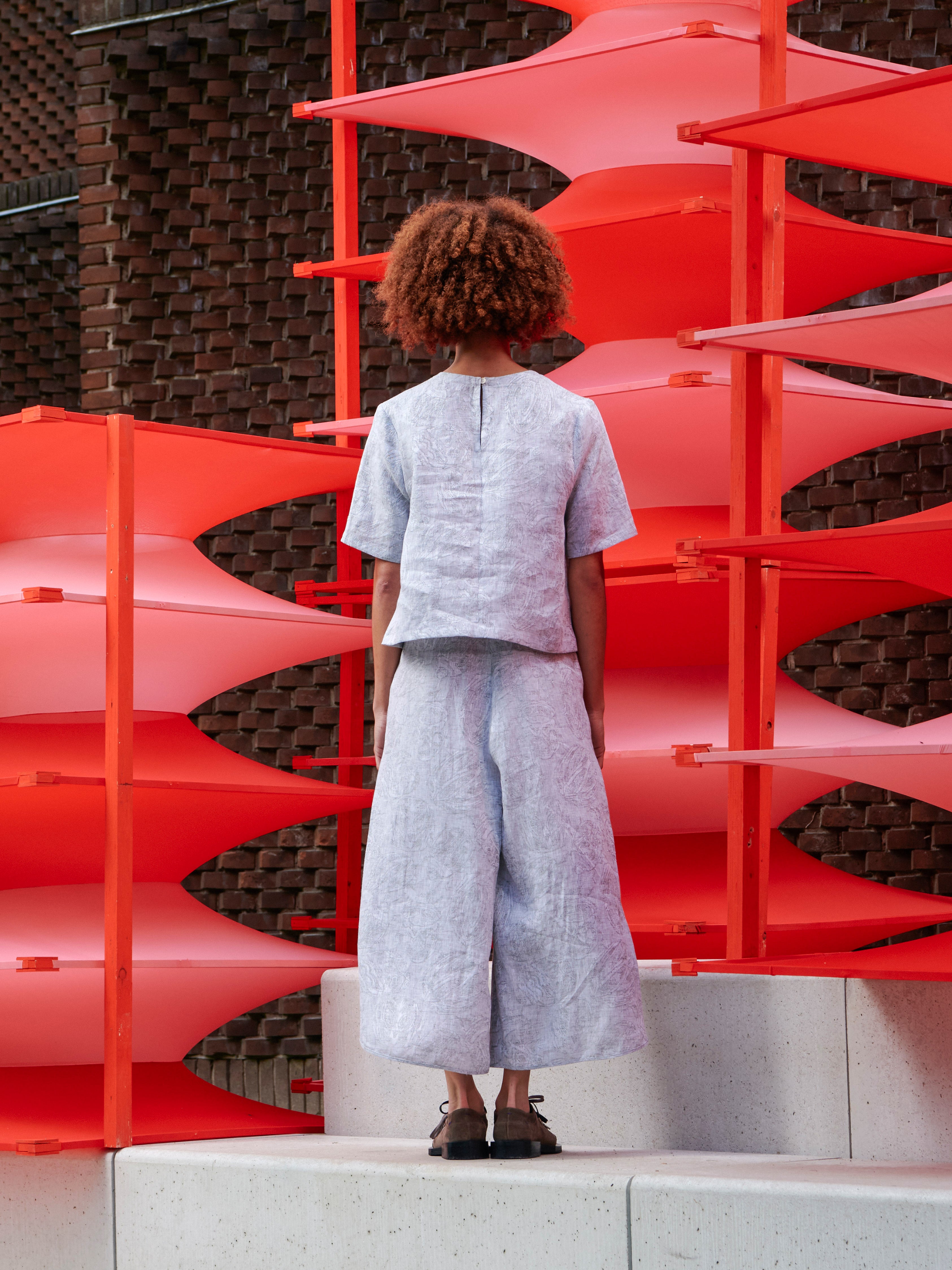 Model wearing the Helmet Top in Bike Helmets grey short sleeve top with button closure at back neck and straight hem, back view – Henrik Vibskov Spring/Summer 2026 Key Collection