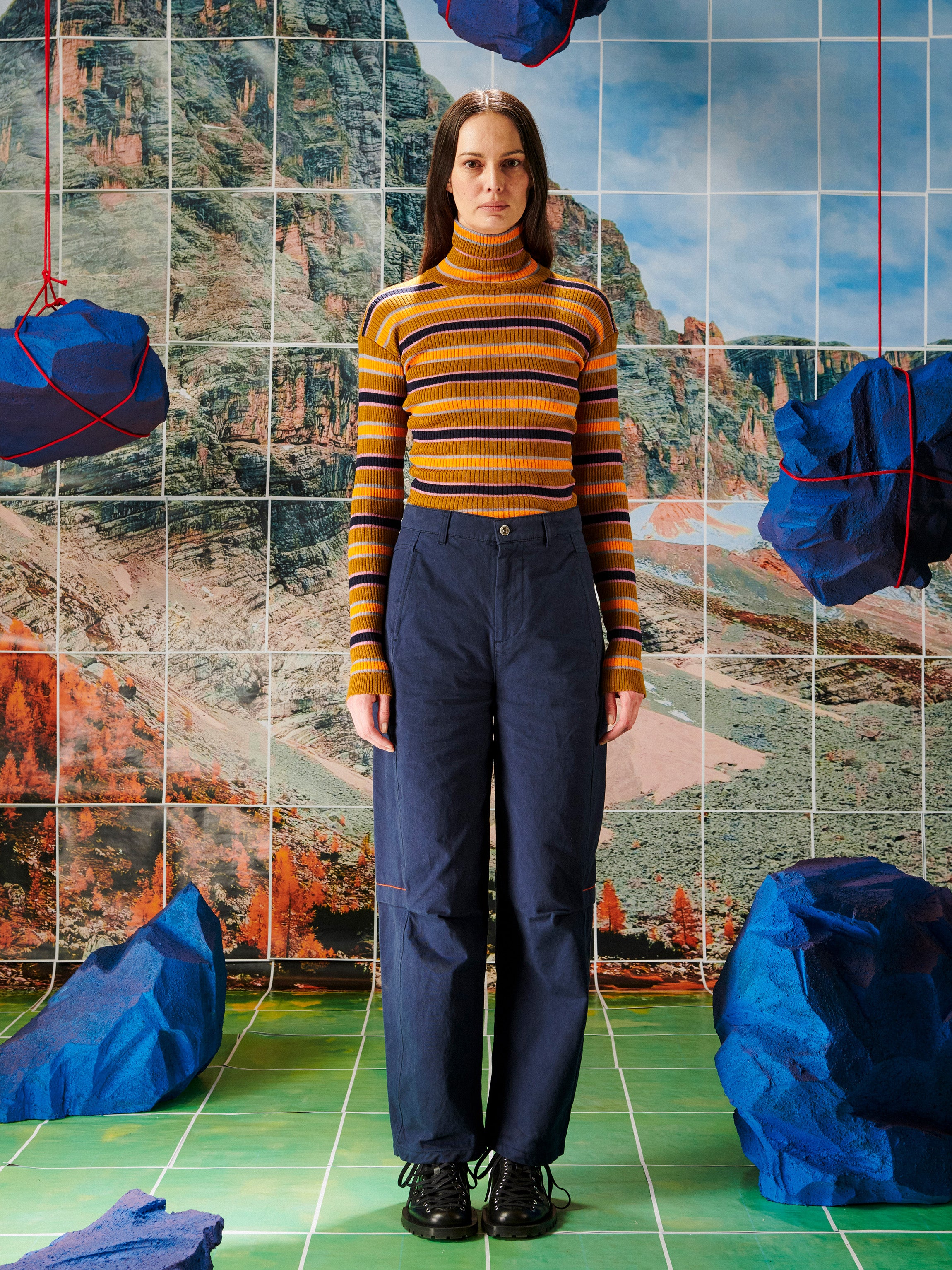 Person wearing a striped sweater and pants standing in front of a collage with mountains and blue hanging rocks.