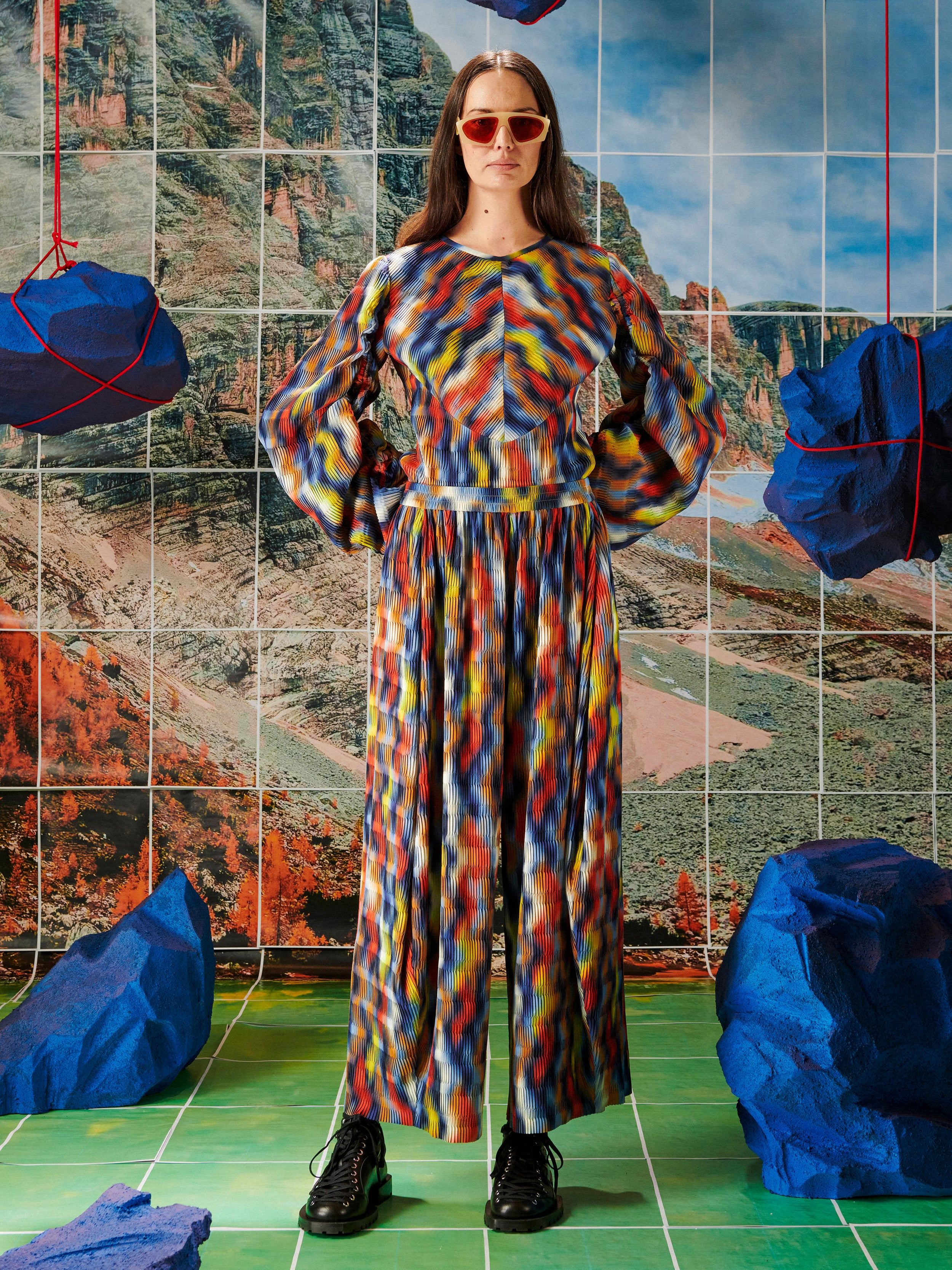 Woman in a colorful pleated top and pants from Henrik Vibskov standing in front of a tiled wall with mountain illustration.