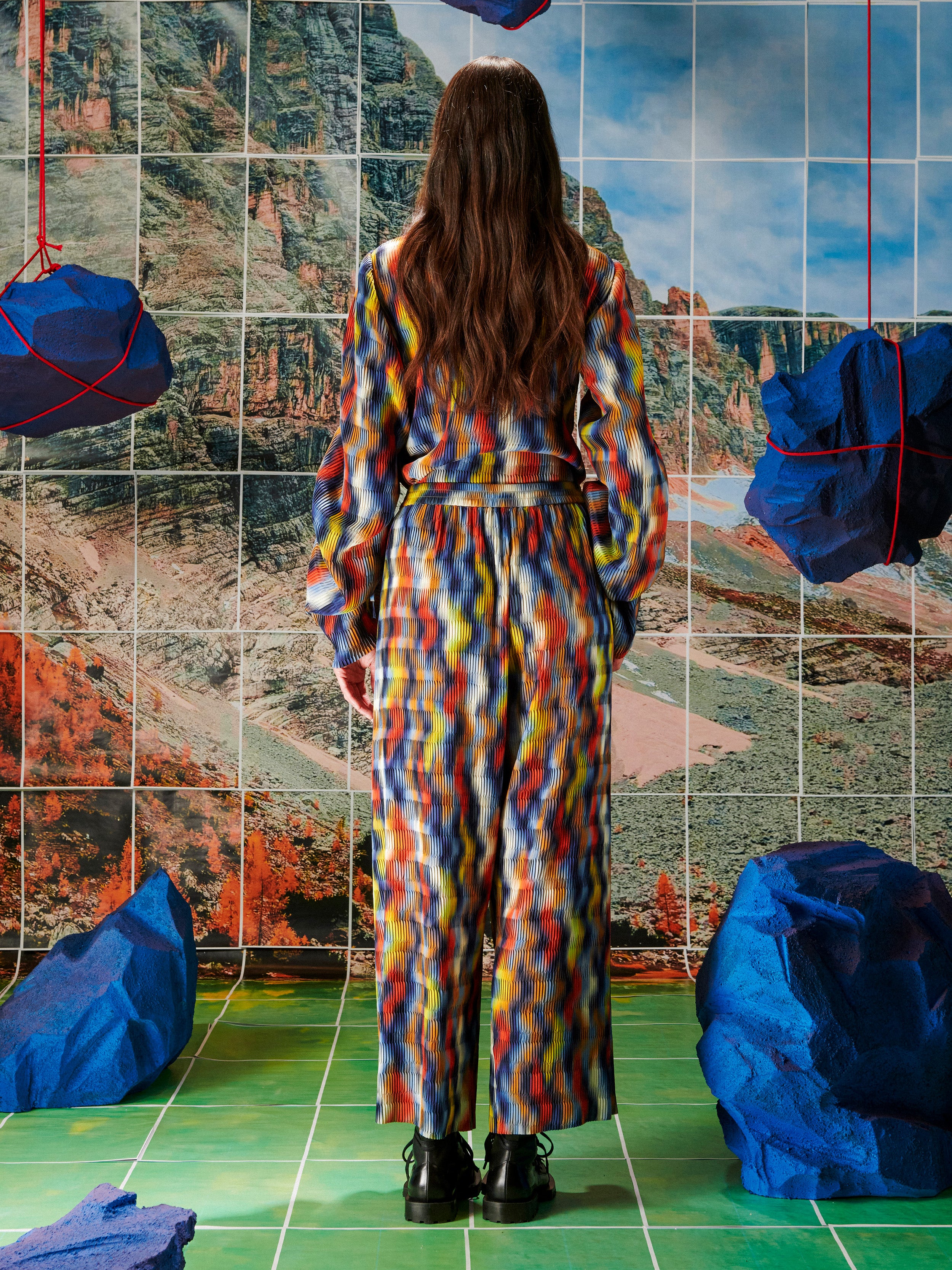 Woman in a colorful pleated top and pants from Henrik Vibskov standing in front of a tiled wall with mountain illustration.