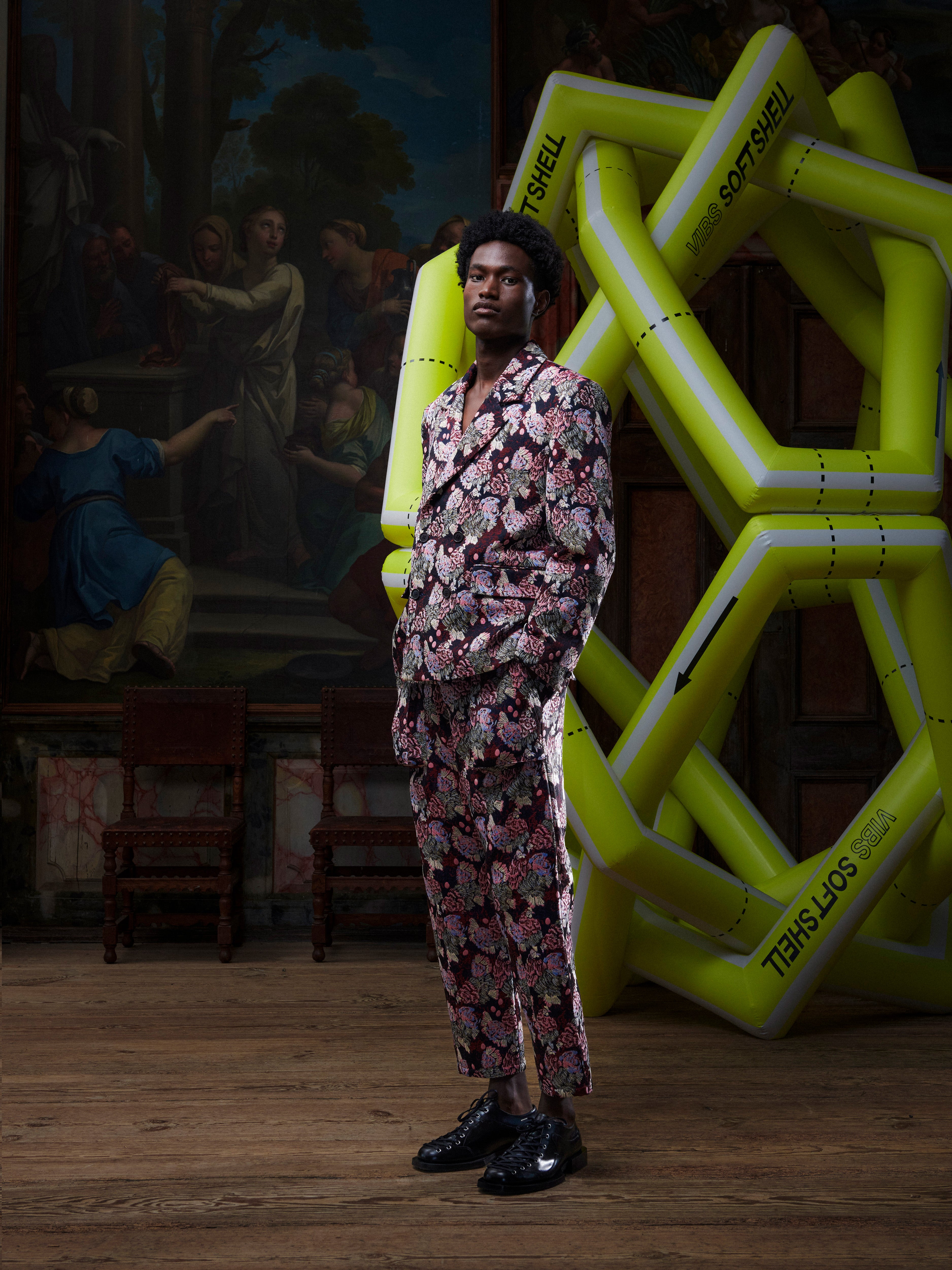 Male model wearing a patterned suit by Henrik Vibskov, standing in front of a large inflatable sculpture with a painting in the background.