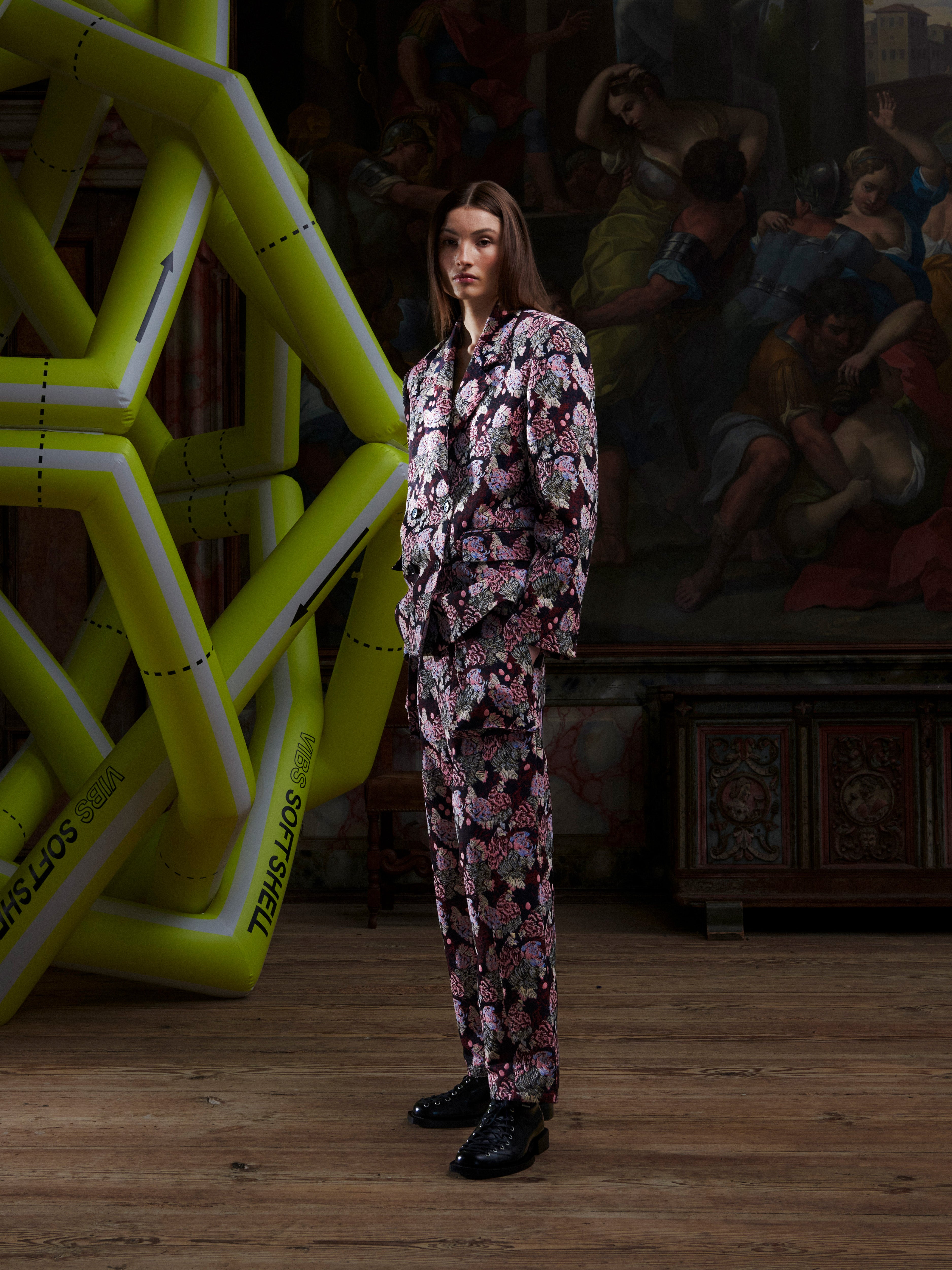 Female model wearing a patterned suit by Henrik Vibskov, standing in front of a large inflatable sculpture with a painting in the background.