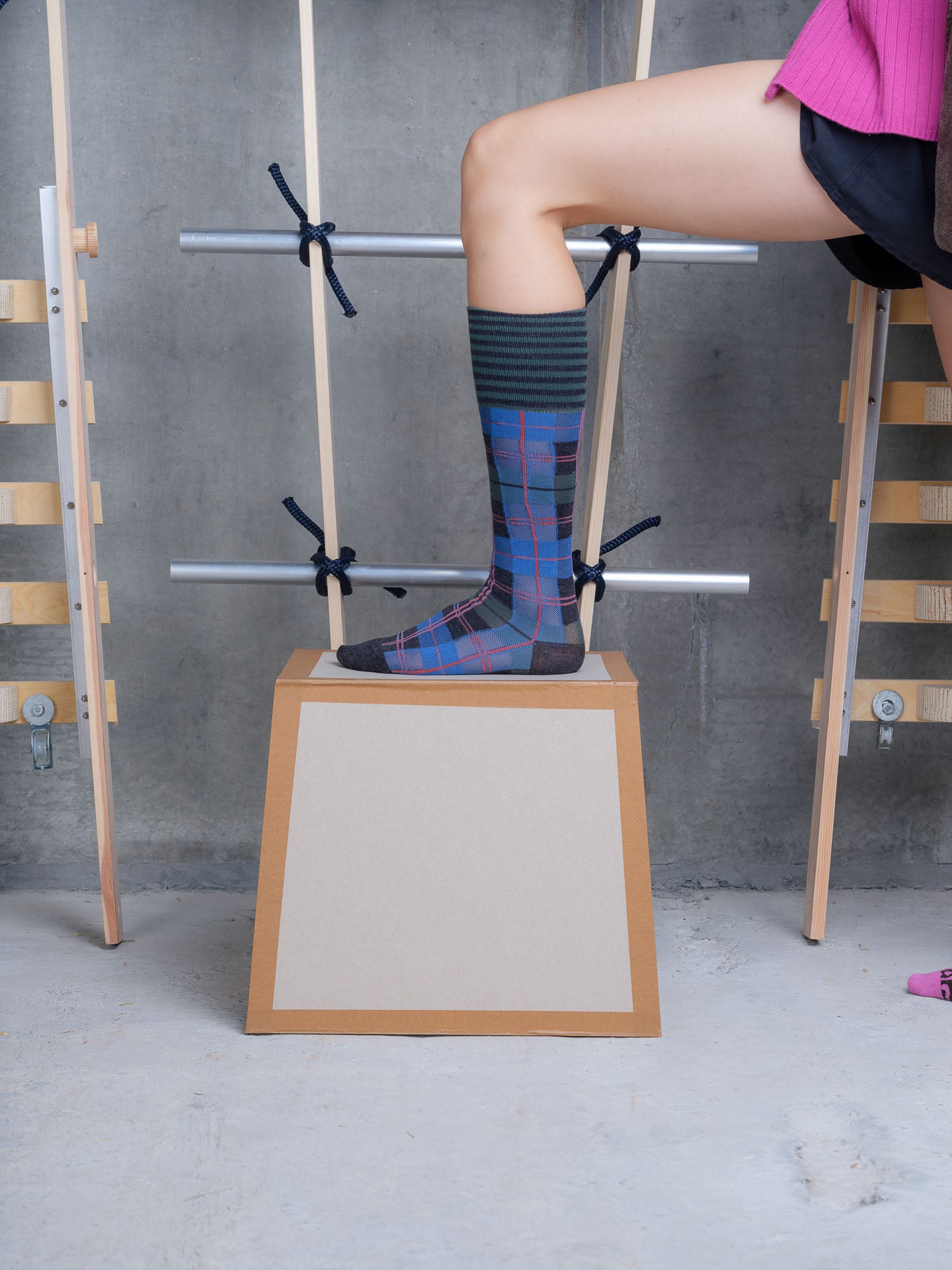 Model wearing Henrik Vibskov Ribbit Knee Sock in blue and green check with dashed red lines, striped cuff and contrast toe