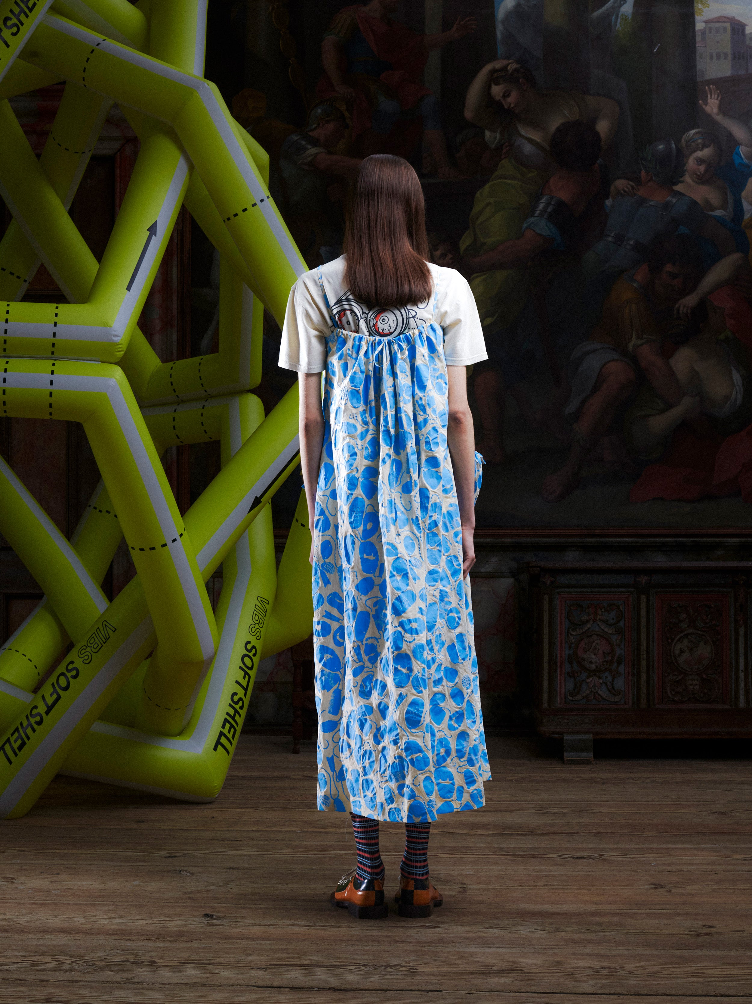 Back view of a woman wearing the off-white and light blue pattern, sleeveless dress on top of a white t-shirt from Henrik Vibskov.