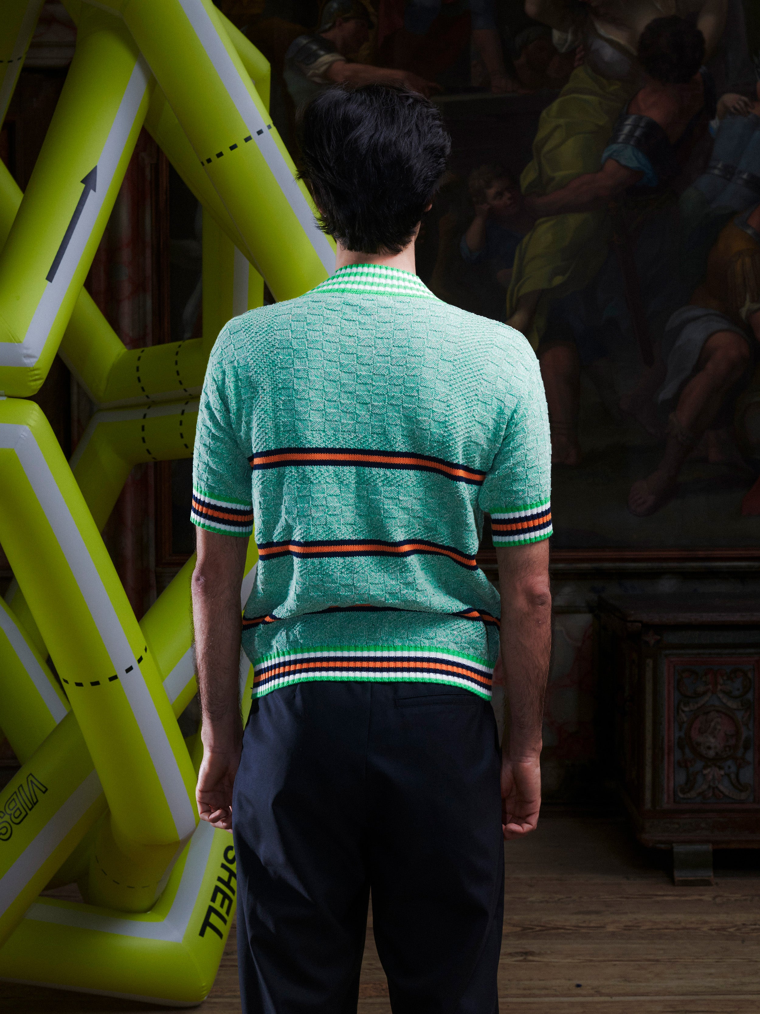 Back view of man wearing the mint green knitted top with stripe details from Henrik Vibskov with a yellow inflatable sculpture in the background.