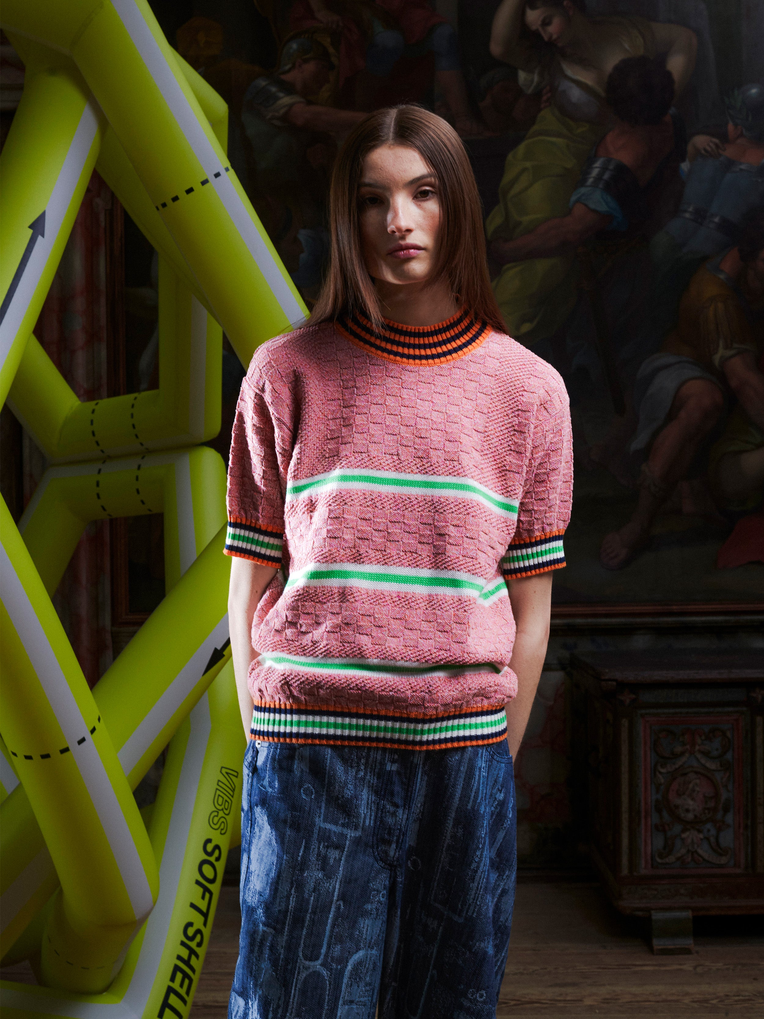 Woman wearing the pink-green knitted top with stripe details from Henrik Vibskov with a yellow inflatable sculpture in the background.