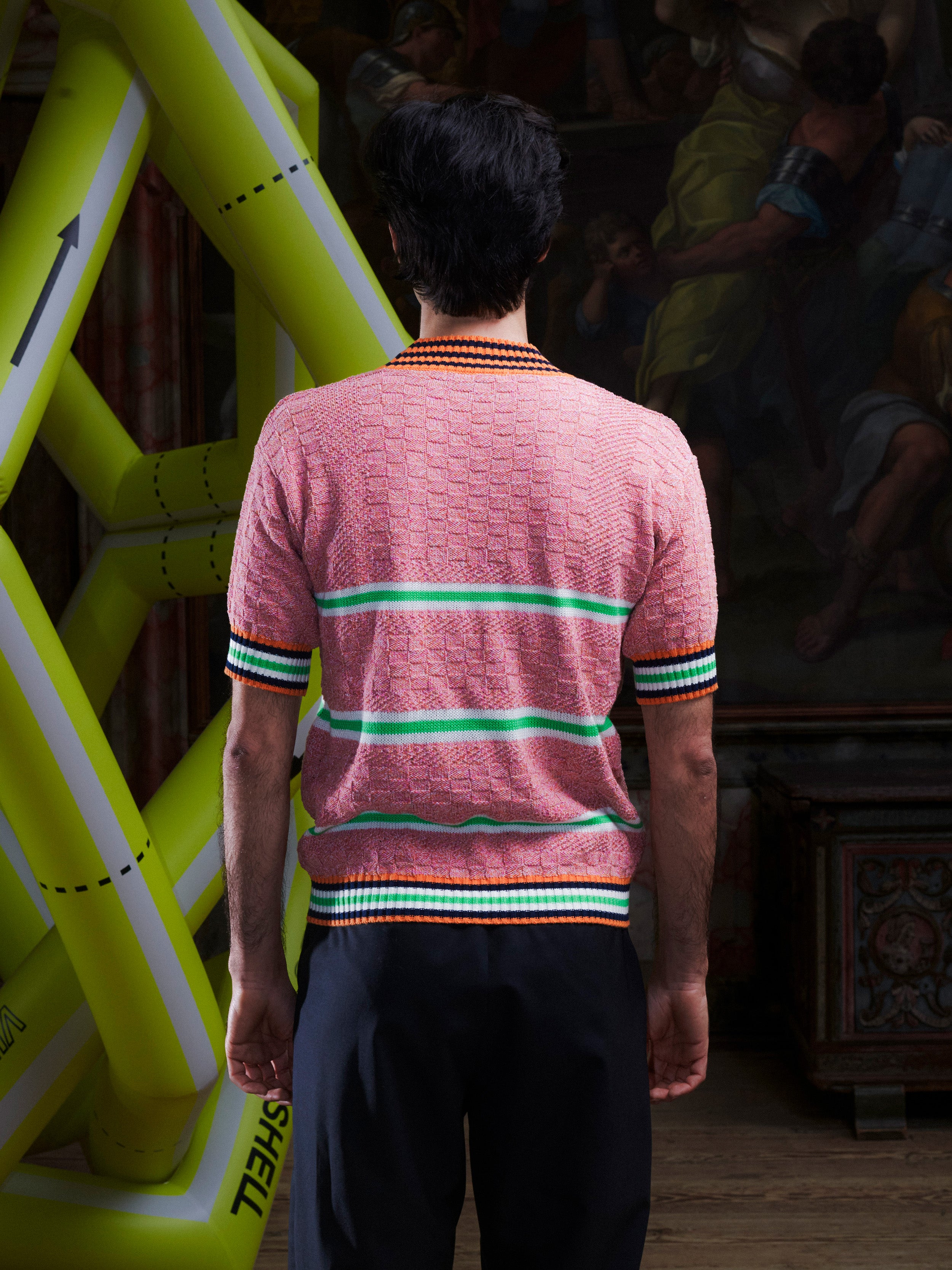 Back view of man wearing the pink-green knitted top with stripe details from Henrik Vibskov with a yellow inflatable sculpture in the background.